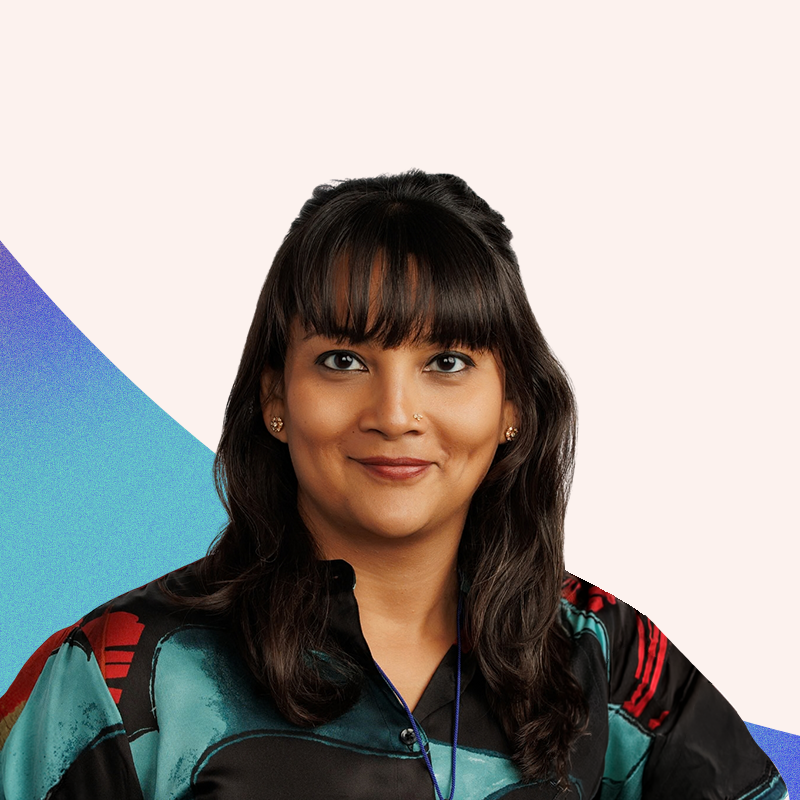 Nayantara Sen - Managing Director, Give Not Take Media - A woman with dark hair, bangs, and a nose piercing, smiling, wearing a colorful patterned shirt, against a background with a white, blue, and purple geometric design.