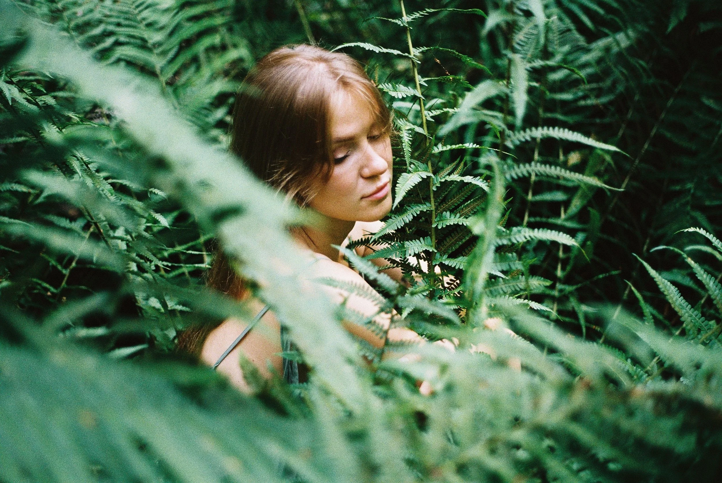 A young woman with long, light brown hair and freckles is among lush green ferns in a dense forest.