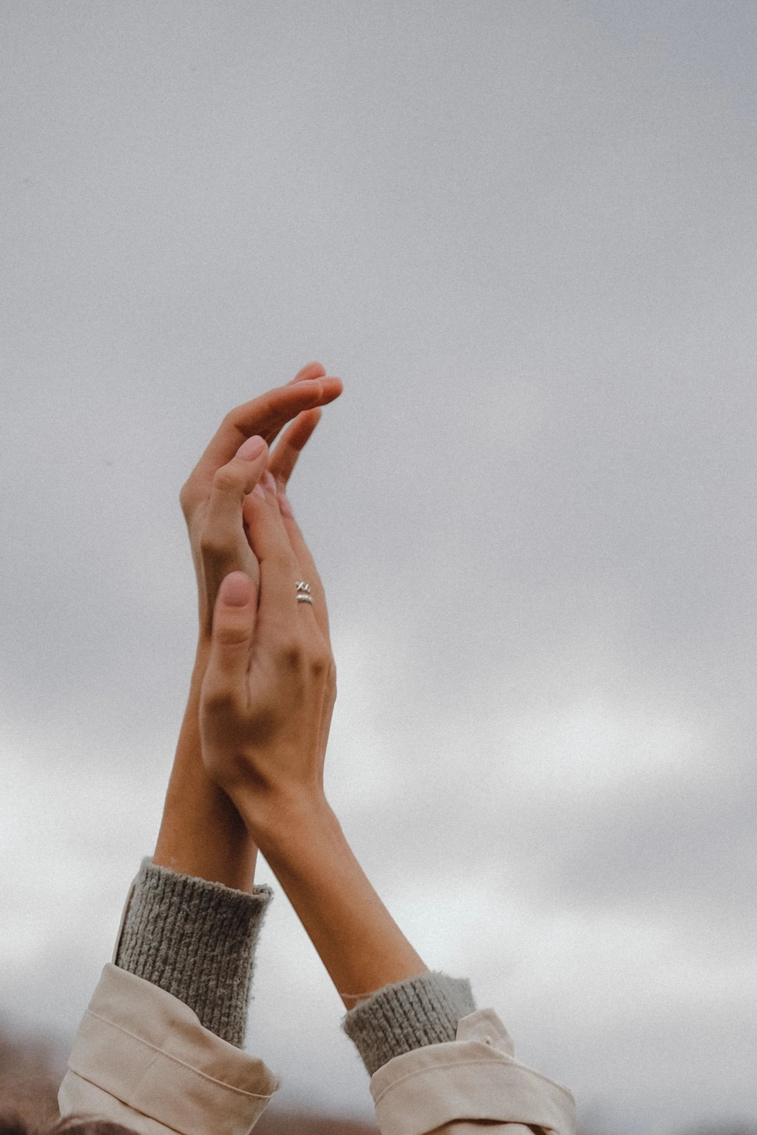 A person's hands with rings, raised against a cloudy sky, with onehand partially overlapping the other. The person is wearing a beige jacket with gray knitted cuffs.