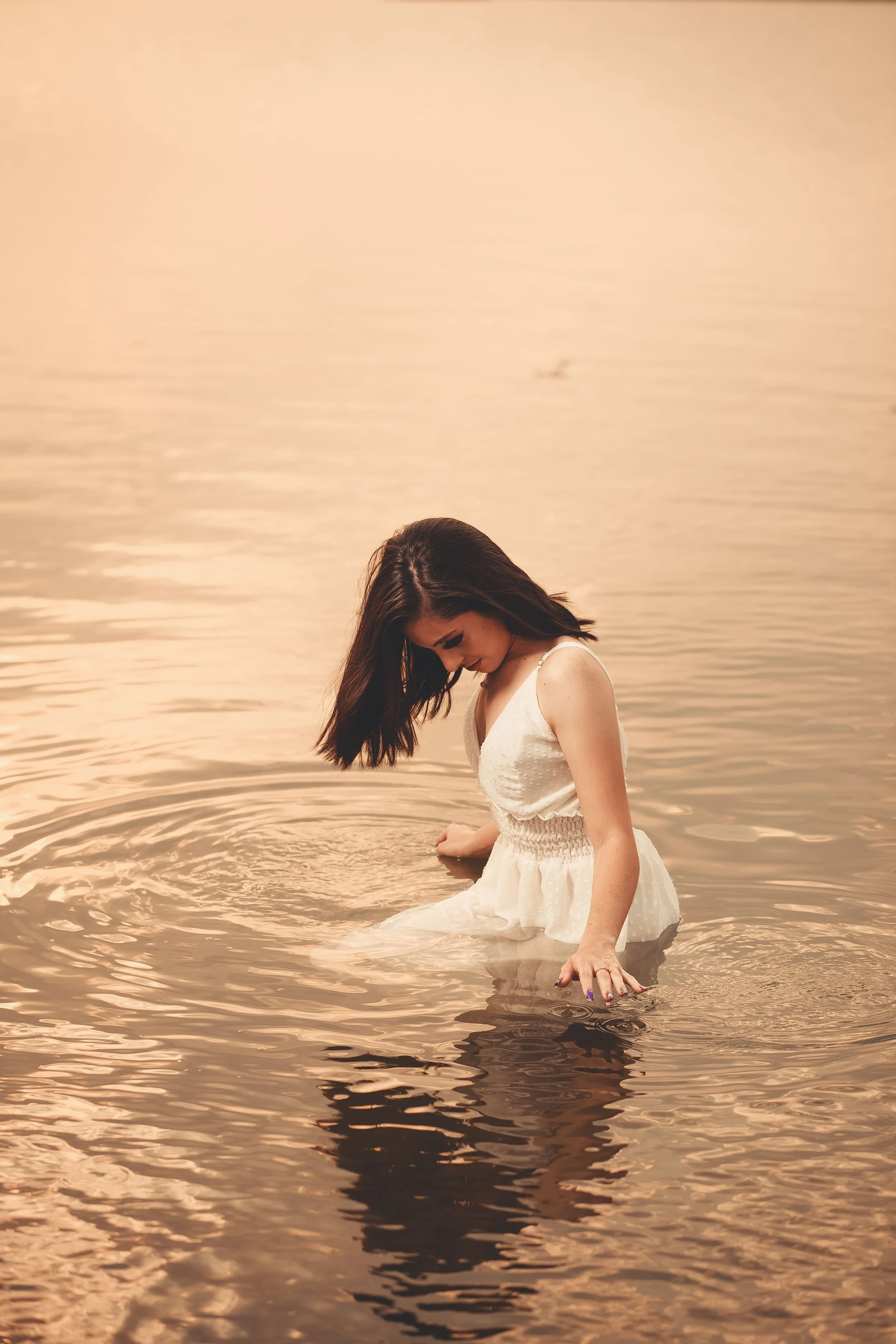 A woman in a white dress sits in shallow water at a lake during sunset, with her head bowed and hair falling over her face, creating a peaceful and reflective scene.