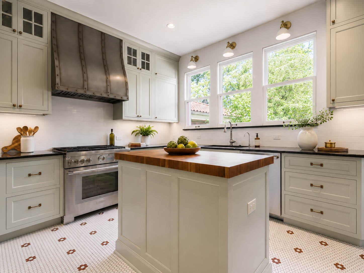 Bright kitchen with pale green cabinets, a stainless steel stove, and a wooden-topped island. Large windows reveal green trees outside. Decorative items include a bowl of green apples, potted plants, and kitchen utensils.