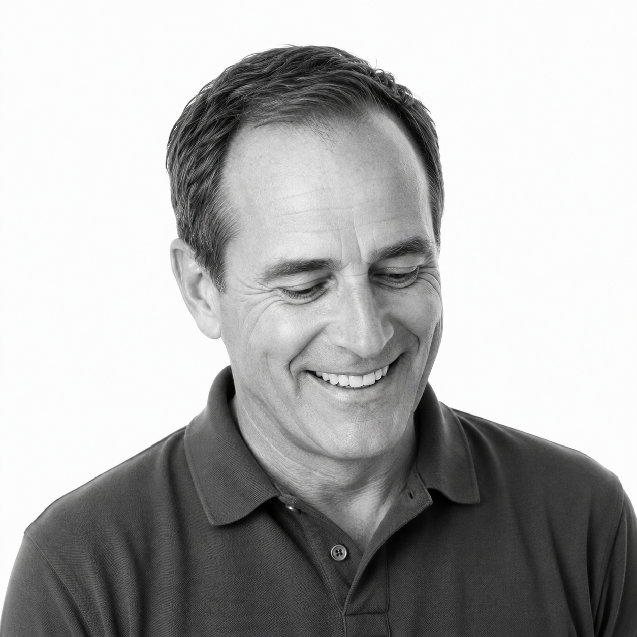 Black-and-white portrait of a man smiling and looking downward, wearing a collared shirt.