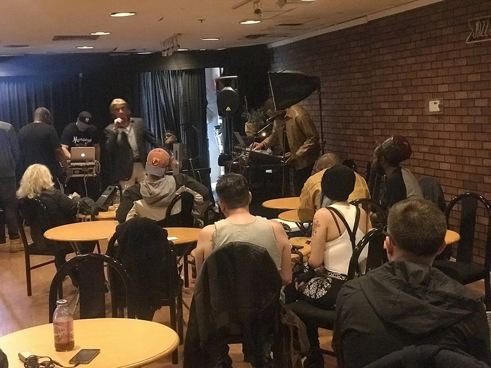 People sitting at tables in a room watching a live music performance with a pianist on stage. The setting appears to be a small venue or cafe with brick walls and dark curtains.