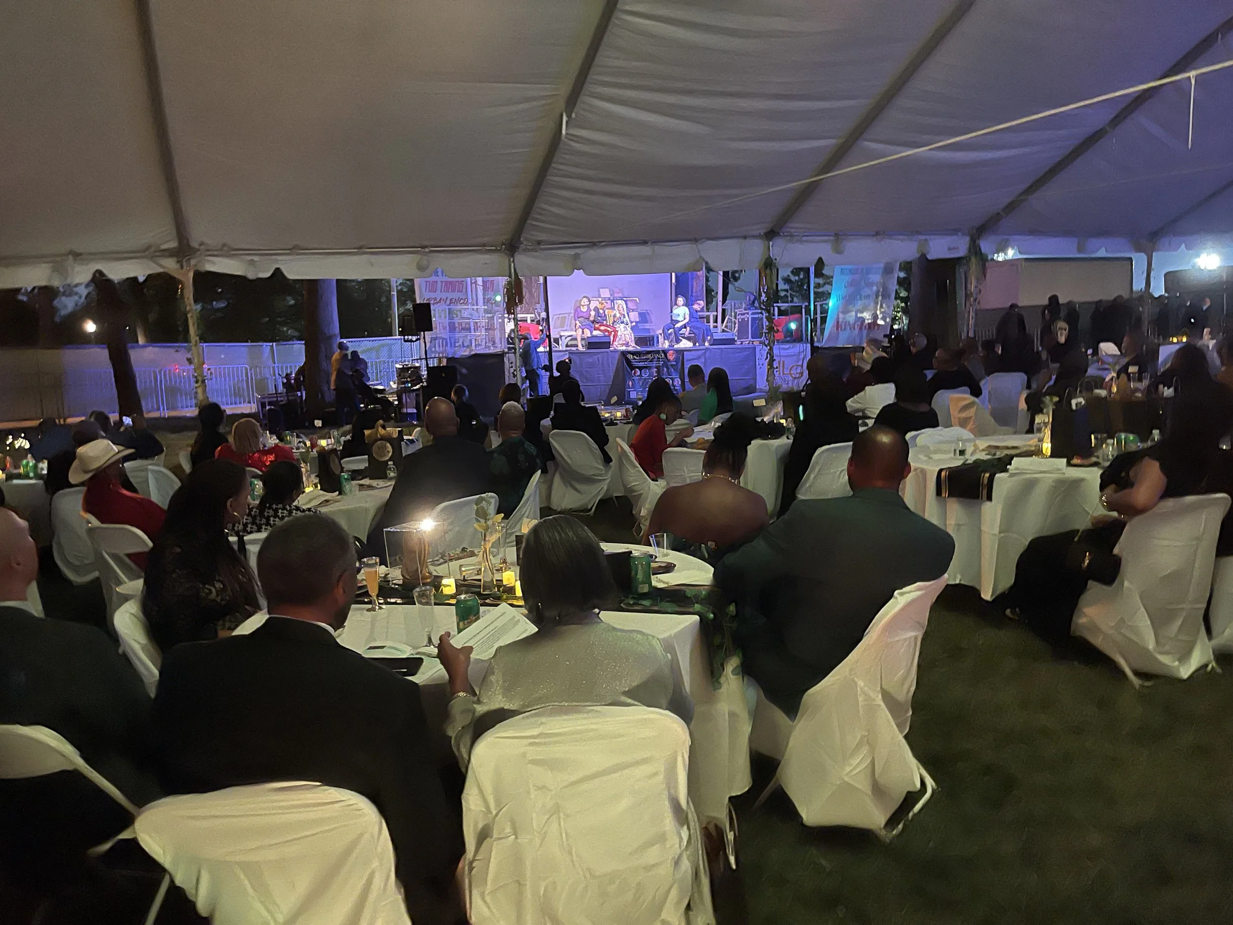 People sitting at tables under a large tent at night, attending a live event with a stage and performers.