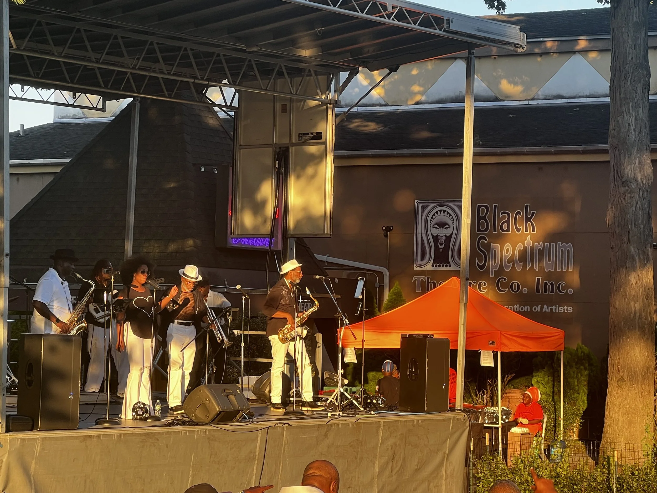 A jazz band performing on outdoor stage at sunset, with musicians playing instruments like saxophone and keyboard, under a metal canopy, with a banner reading 'Black Specturm' and some audience members including a woman in red sitting near a large tree.
