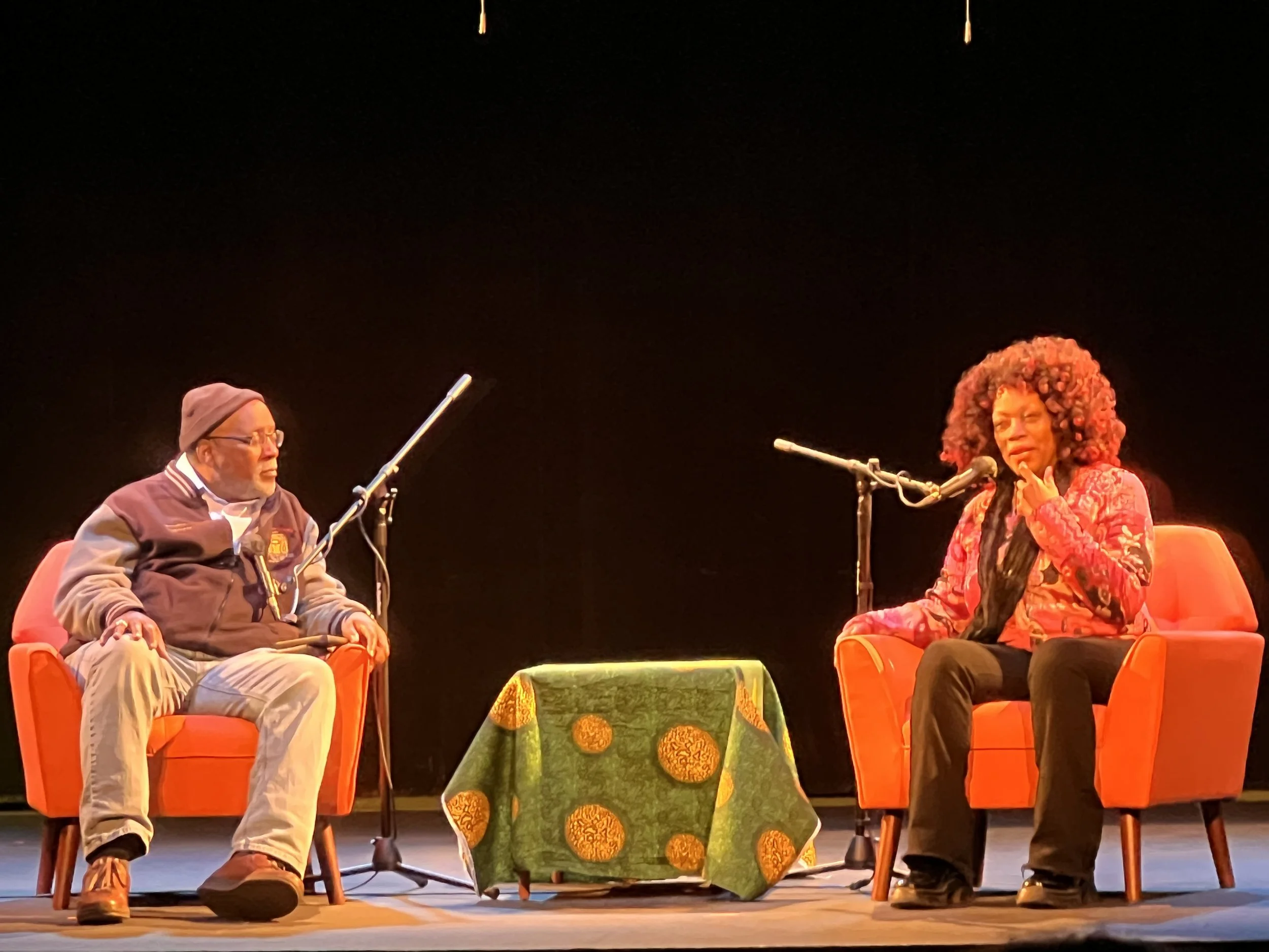 Two people sitting on orange chairs on a stage, engaged in a discussion or interview, with a small table covered with a green patterned cloth between them.
