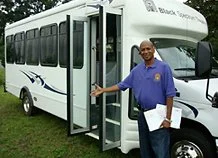 A smiling man in a blue shirt standing at the entrance of a white shuttle bus with green and blue accents, parked outdoors on grass.