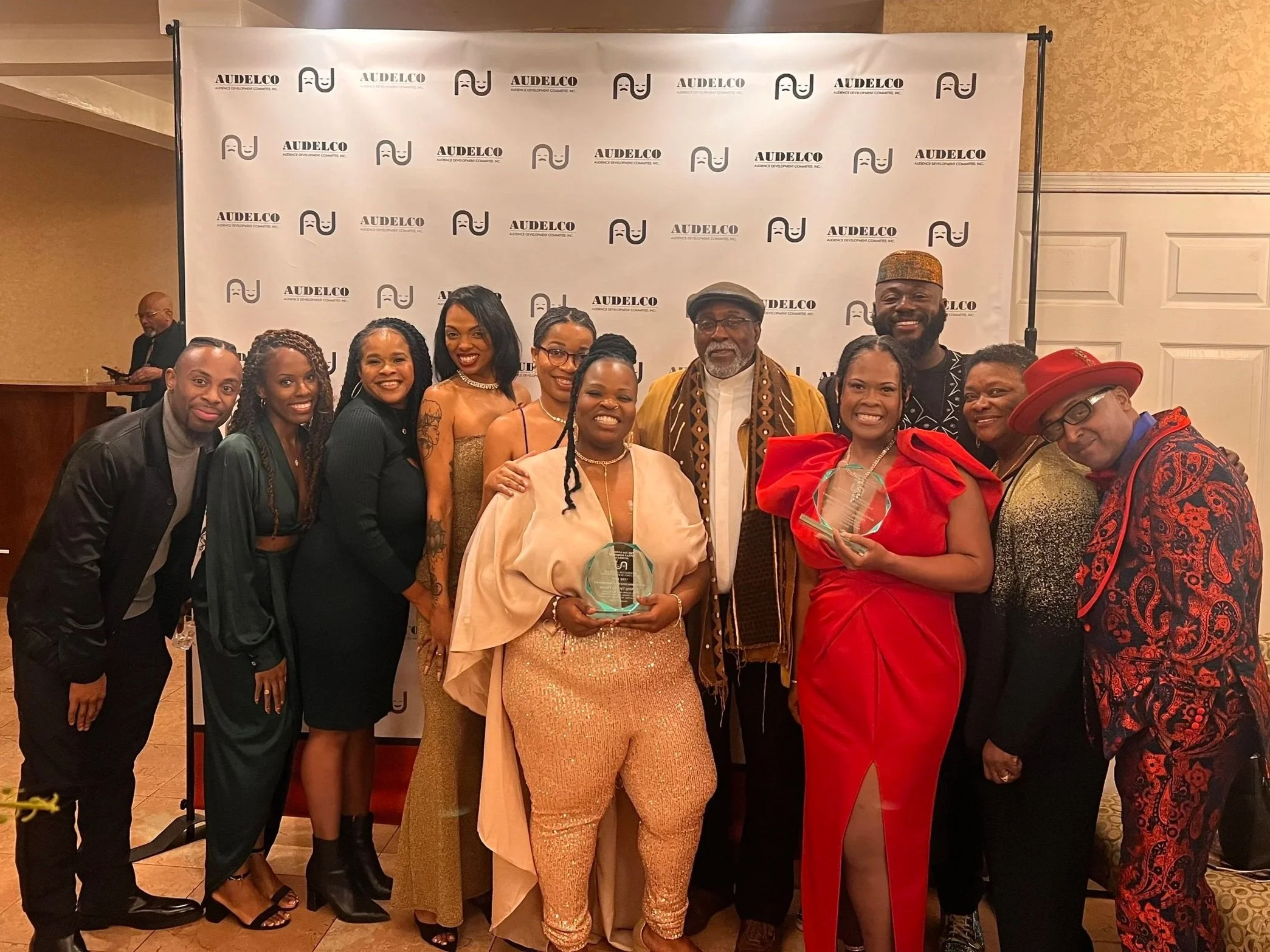 Group of people at an event posing for a photo in front of a backdrop with the AUDELCO logo, with some individuals holding awards.