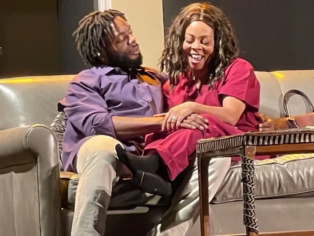 A man with dreadlocks and a woman with curly hair sitting closely on a couch, smiling and holding hands. The woman is dressed in a red outfit, and the man is in a purple shirt and beige pants. There is a small table in front of them.