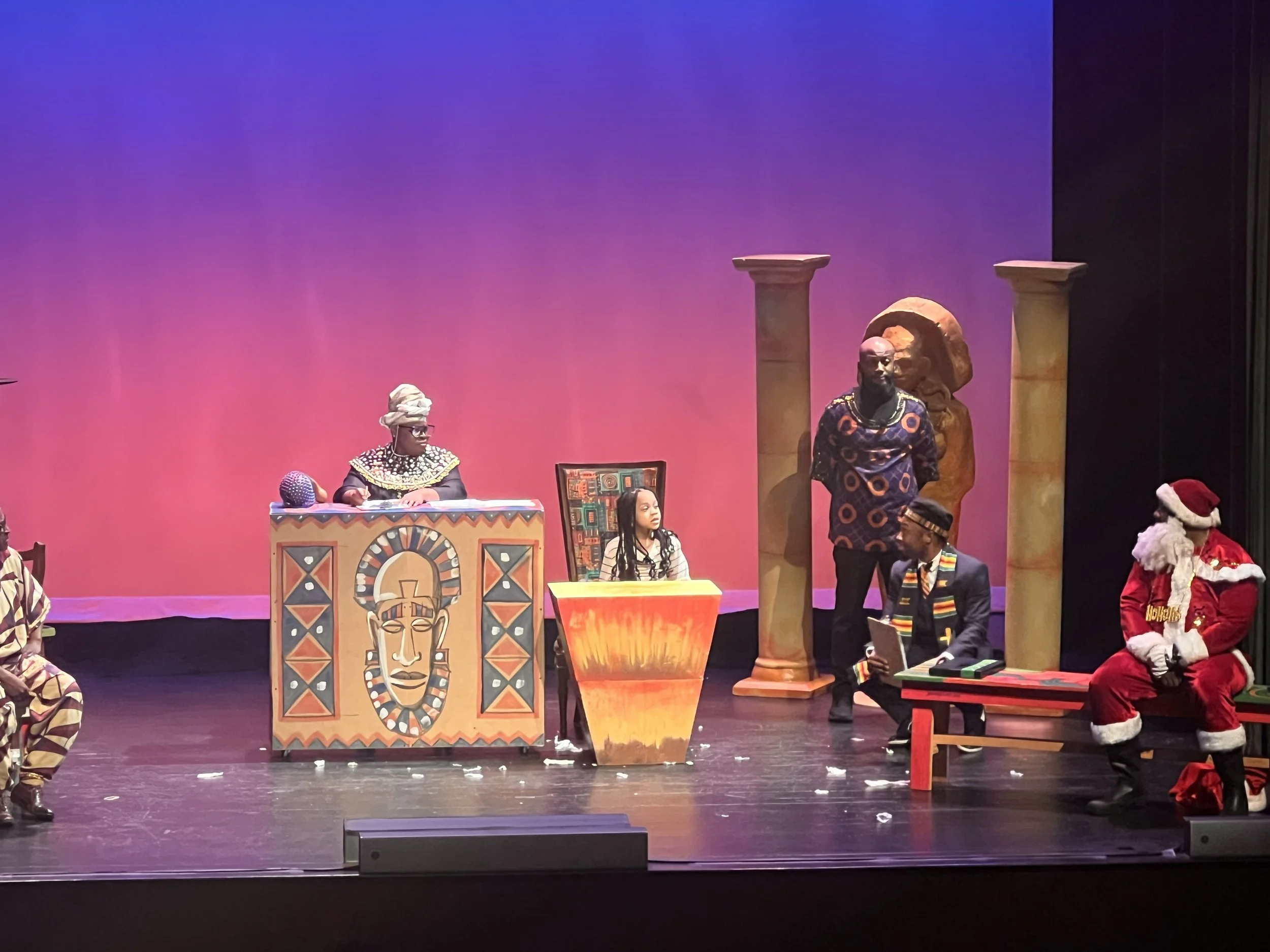 On stage, performers wearing colorful costumes with African patterns, including a woman with glasses and a headwrap, a man in a circular patterned shirt, and others dressed as Santa Claus and in traditional attire. The stage has African cultural decorations, statues, columns, and a pink and purple gradient backdrop.