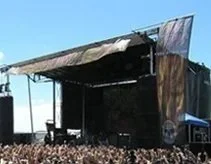 An outdoor music stage with a large overhang, set up at a festival or concert, with a crowd gathered in front.