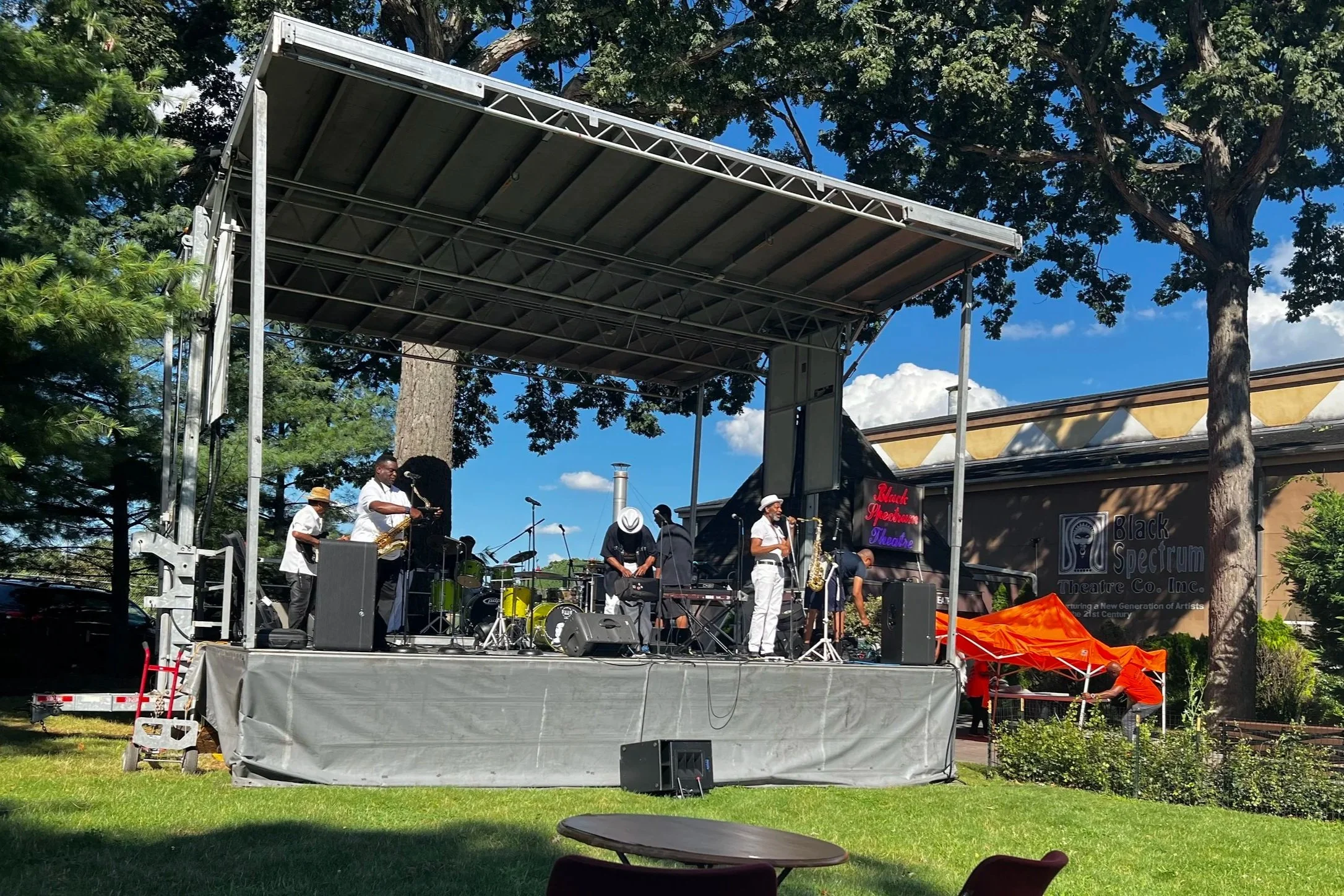 A live outdoor jazz concert on a stage with musicians playing instruments, including saxophones and drums, under a metal canopy. Trees and a building are in the background, with a partly cloudy sky overhead.