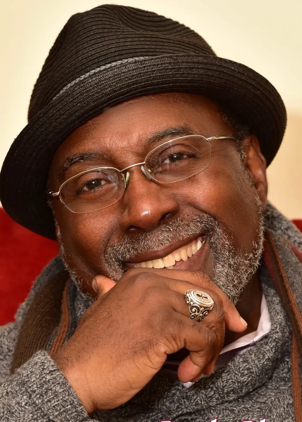 A smiling man wearing glasses, a dark fedora hat, and a gray sweater with a white shirt underneath. He has a gray beard, a silver ring on his finger, and is resting his chin on his hand.