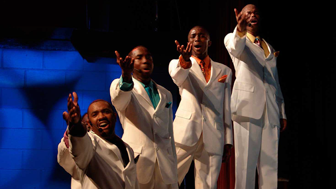 Four male performers on stage, dressed in white suits, singing or acting with expressive gestures, under stage lighting.