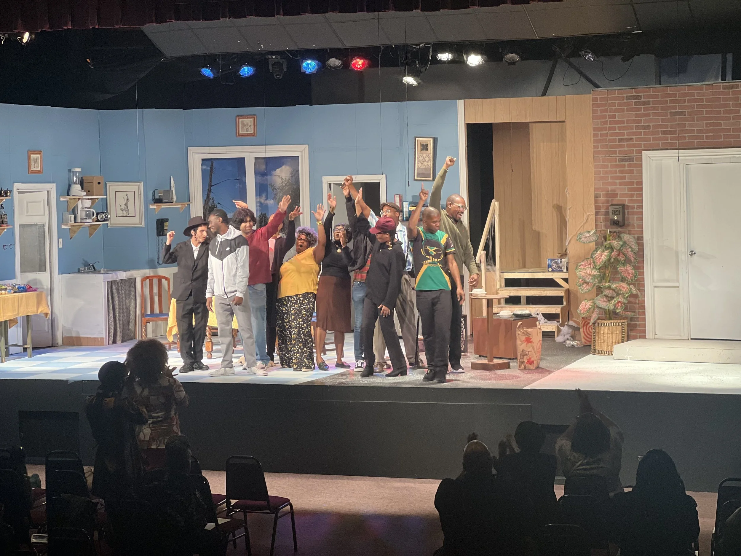 A group of diverse performers on a stage, celebrating with raised hands and smiles, set against a backdrop of a blue wall, windows, and stage scenery in what appears to be a theater or performance venue.