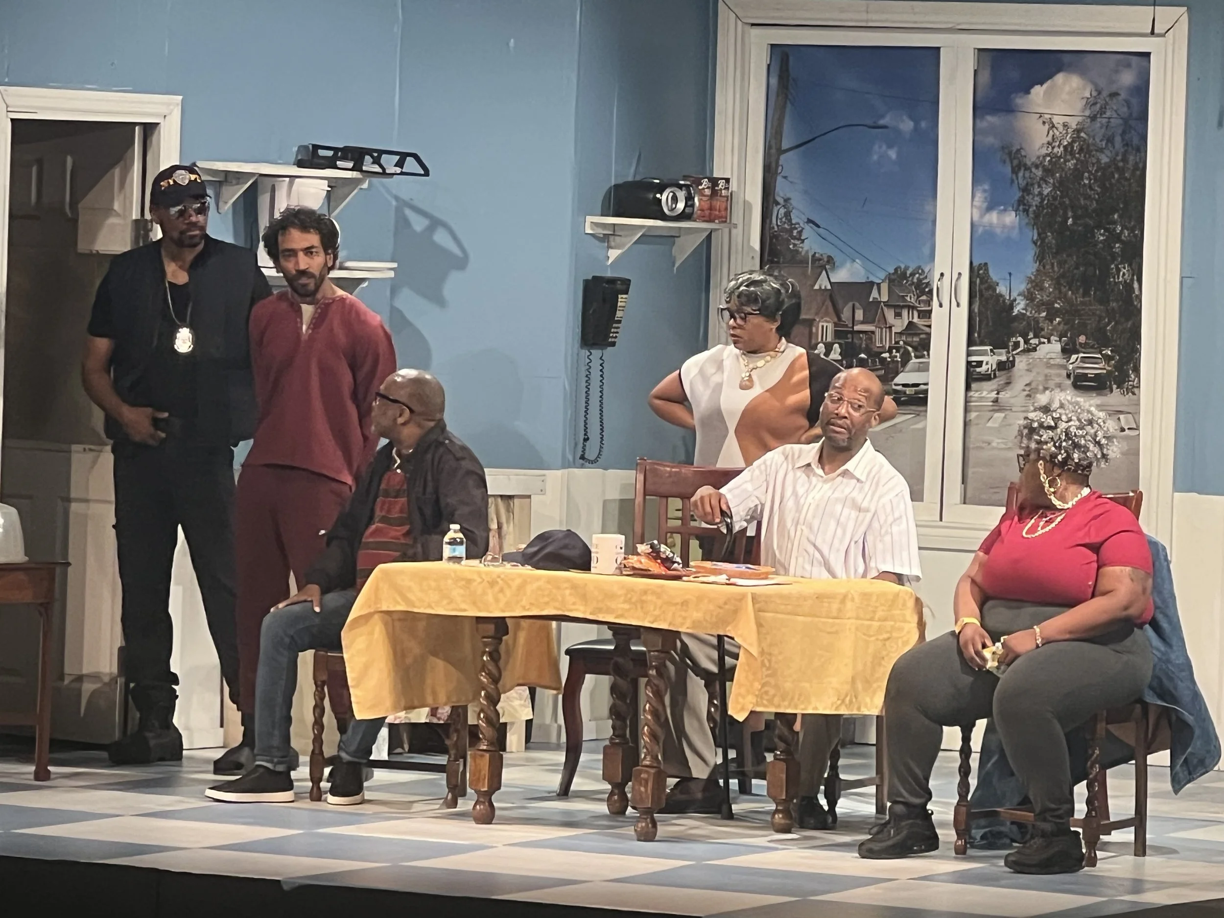 Scene from a play, featuring actors in period costumes sitting and standing around a table with a yellow tablecloth, set in a room with a large window showing a street scene.