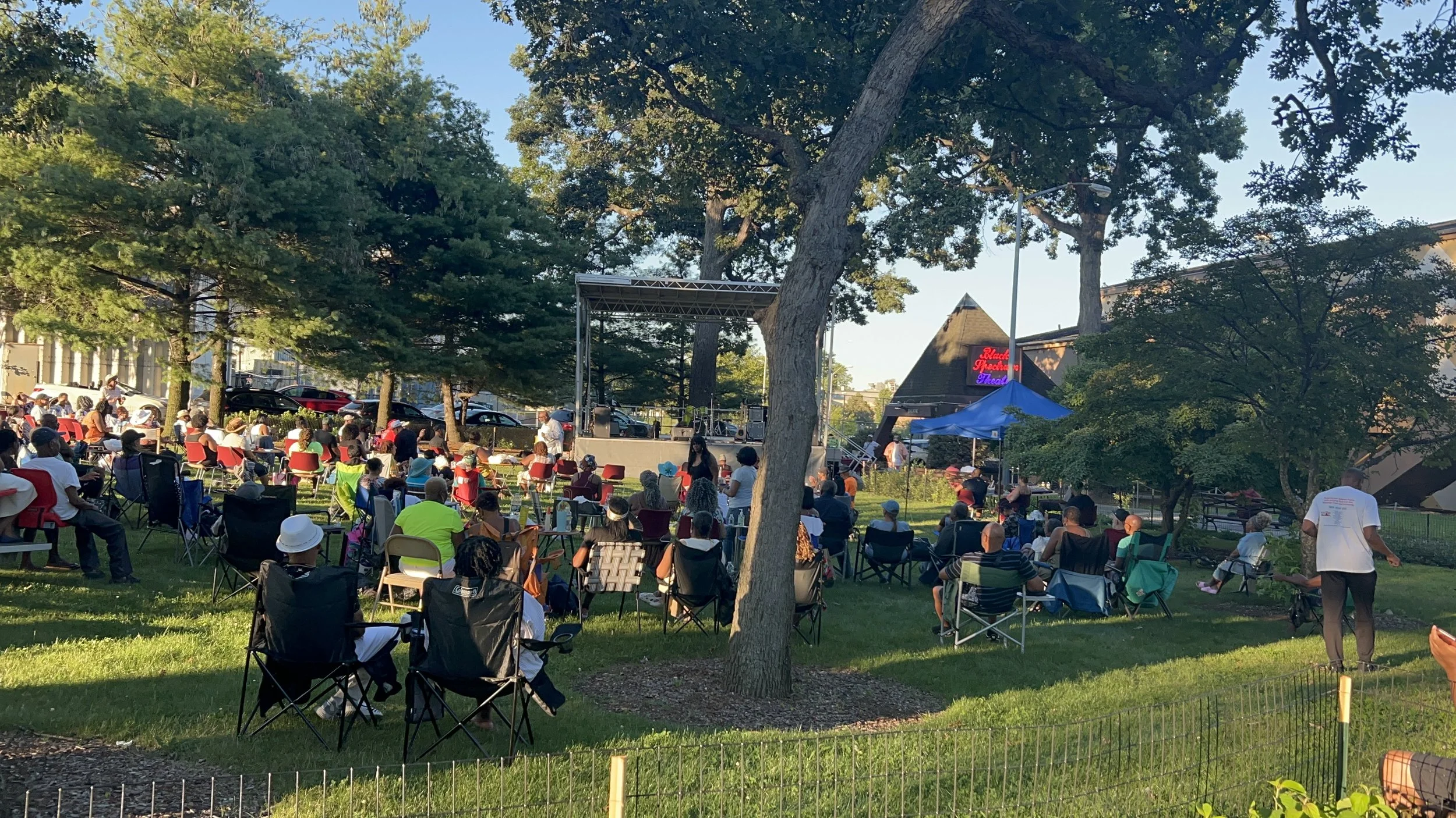 Outdoor concert in a park with many seated and standing attendees, stage with performers, surrounded by trees and sunlight