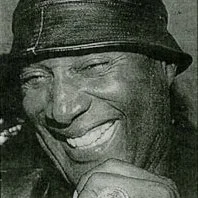 A black and white photo of a man smiling or laughing, wearing a hat.