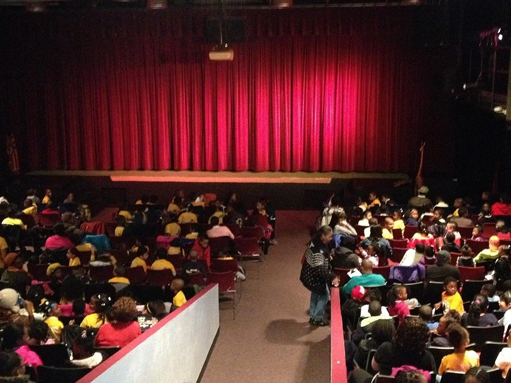 Theater stage with closed red curtains and an audience of children and adults seated in rows.