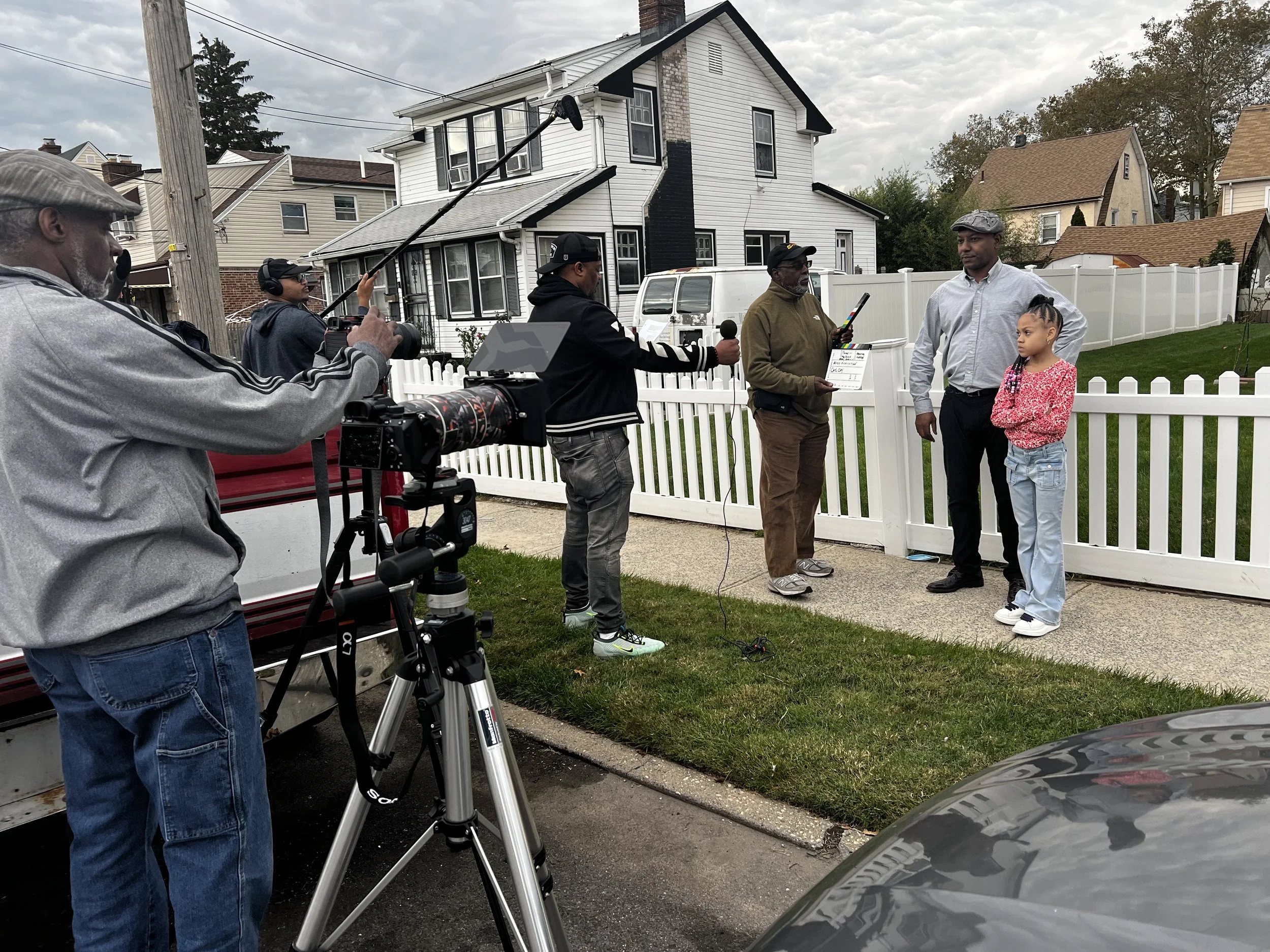 A behind-the-scenes look at a film or video shoot outside a white house. Crew members are filming a man and a girl in front of a white picket fence, with equipment such as a camera, boom microphone, and clapperboard visible.