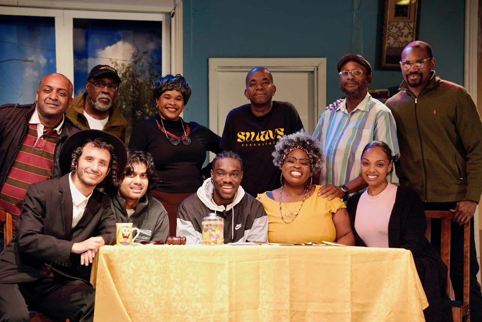 Group of twelve diverse people gathered around a table, some sitting and some standing, indoors with a window showing trees and sky in the background.