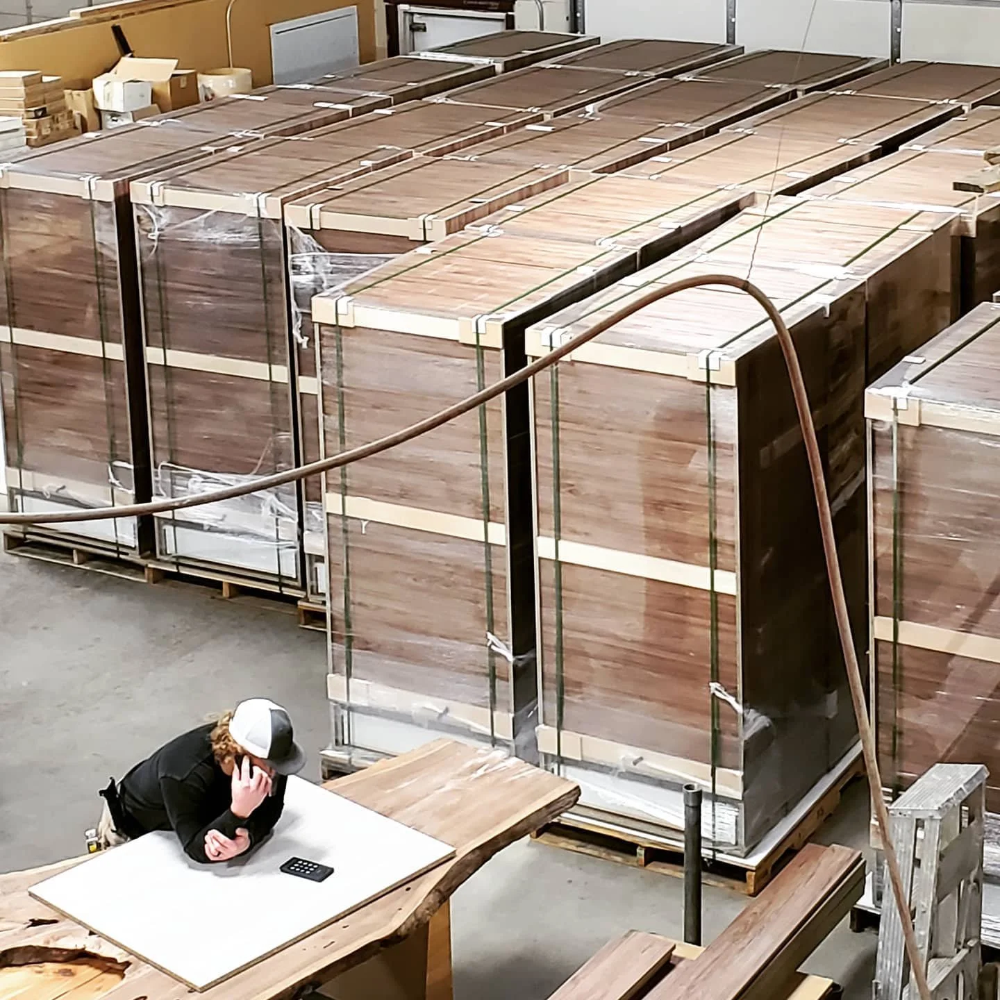 Large crates of wood in a warehouse, with a person in a black jacket and cap sitting at a table talking on a phone.
