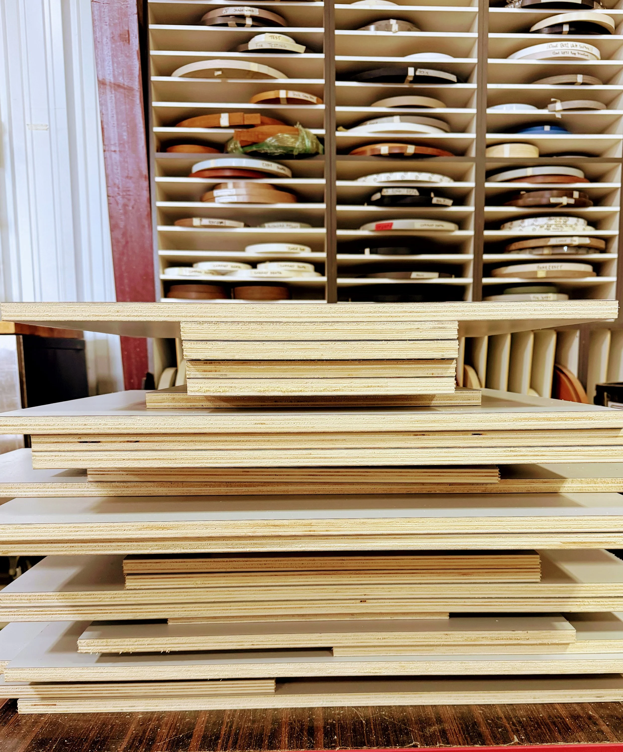Shelf with circular pieces of wood or veneers, each labeled with different names, on a storage rack in woodworking or cabinetry shop.