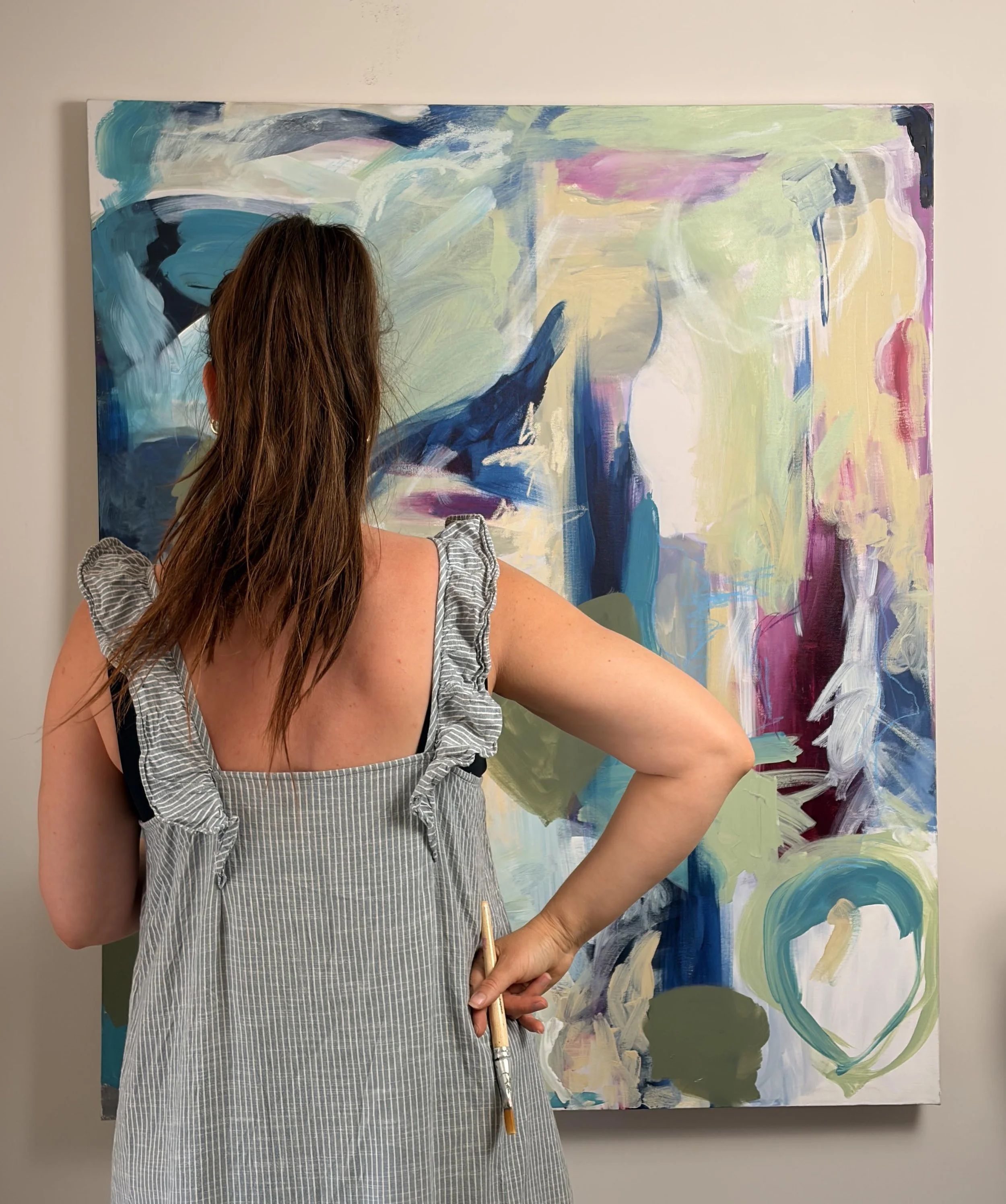 A woman with long brown hair wearing a gray and white striped dress is standing in front of a large abstract colorful painting, holding a paintbrush in her right hand.