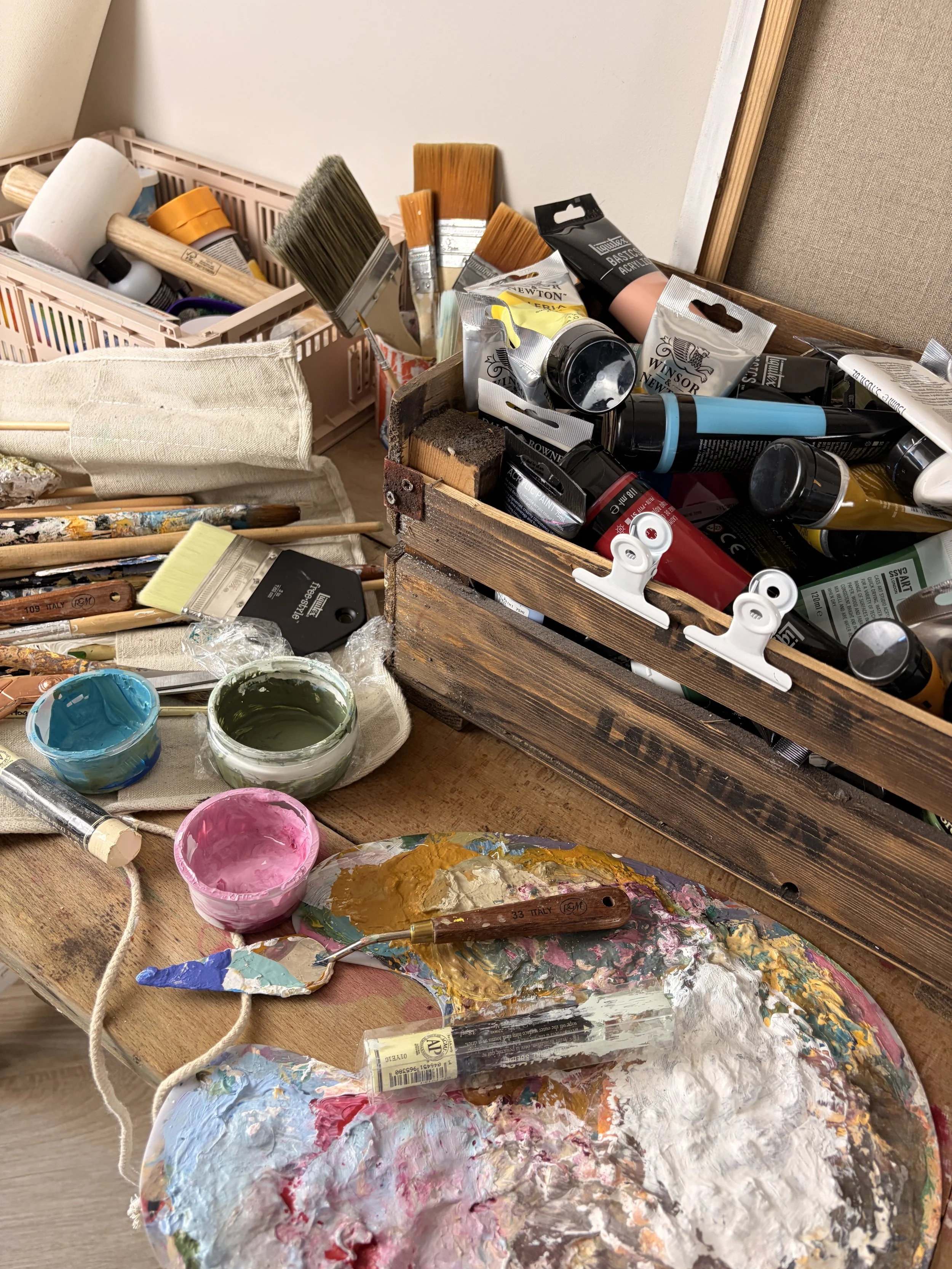 Paint palette with mixed colors, YouTube glue, brushes, paint tubes, and jars of paint on a wooden surface with an open wooden box of art supplies.