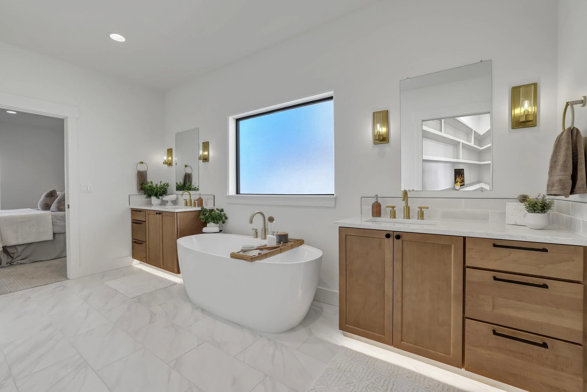 Modern bathroom with a freestanding bathtub, wooden vanity with double sinks, large mirror, and wall-mounted gold light fixtures, next to a window with frosted glass, and a view into a bedroom with a bed.
