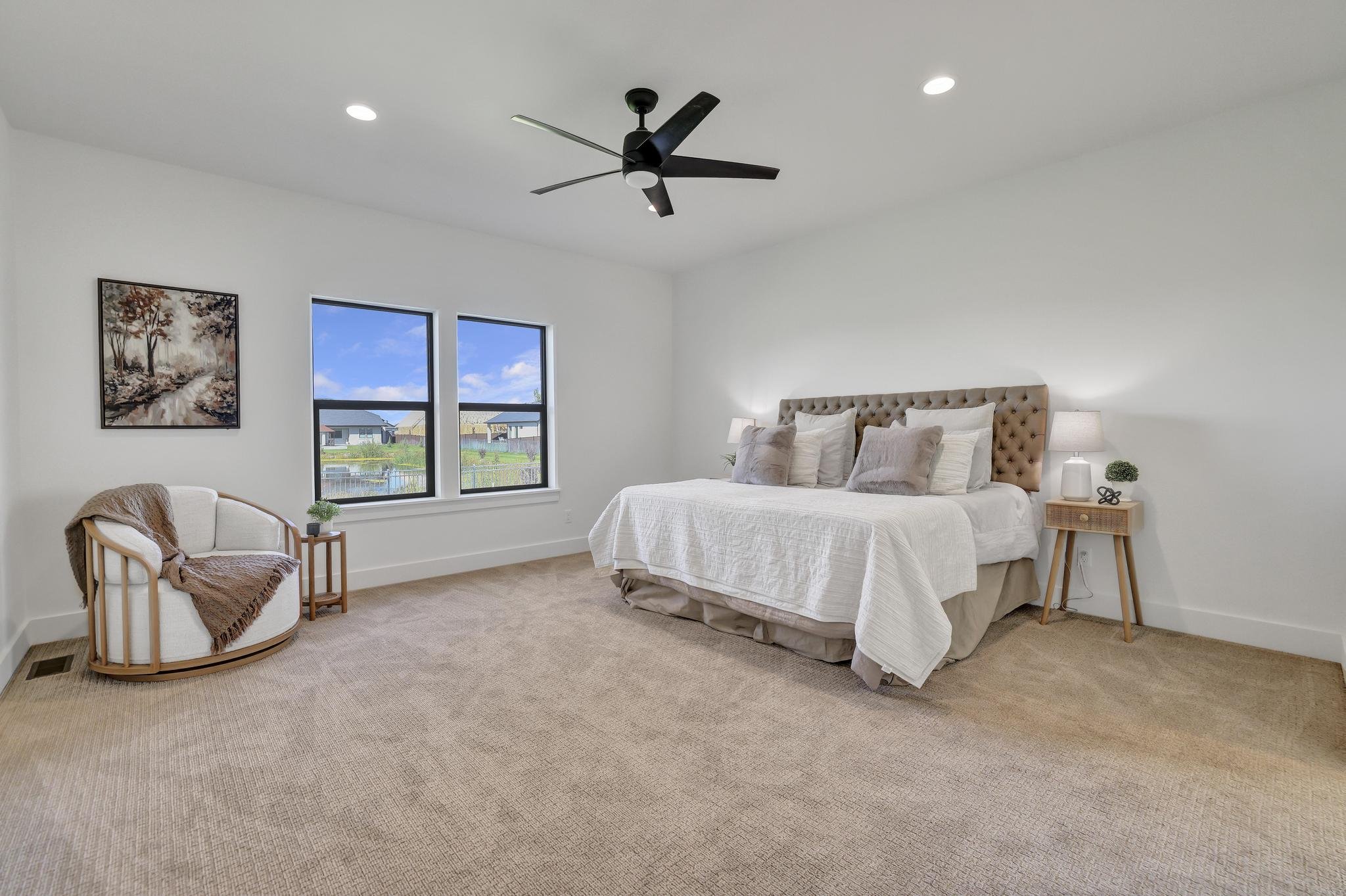A bedroom with a large bed, beige headboard, and white bedding. Two nightstands with lamps, a beige armchair with a brown throw blanket, and artwork of trees hanging on the white wall. Two windows showing a view of neighboring houses and a blue sky.
