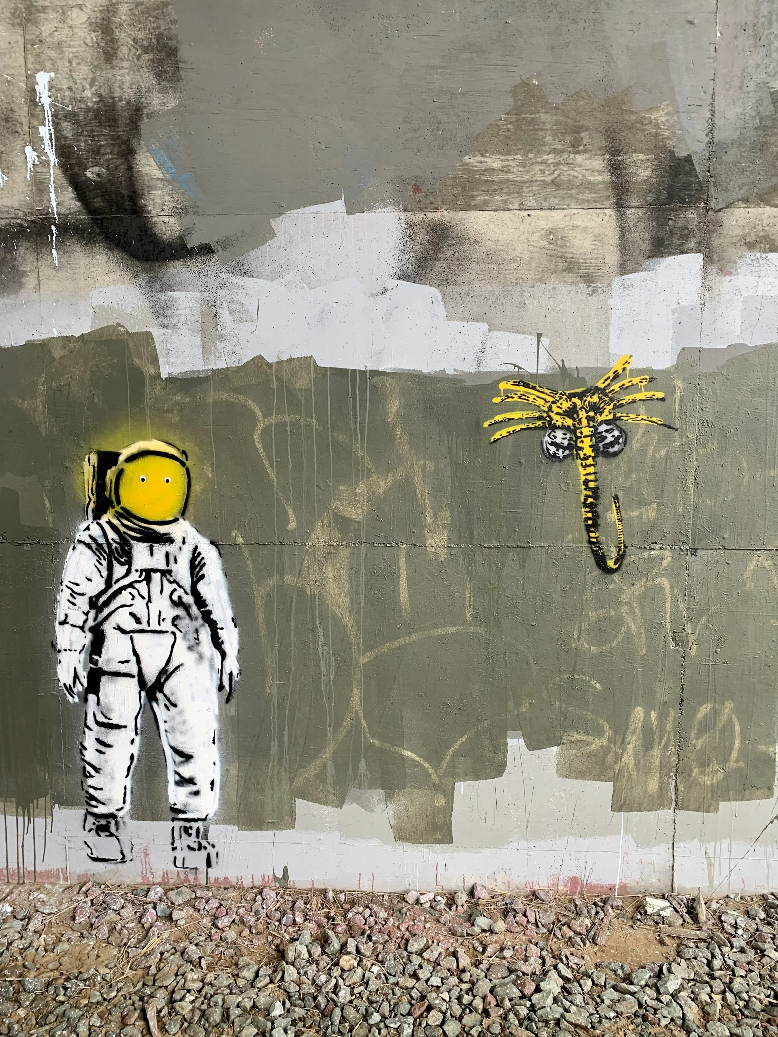 Street art graffiti of an astronaut figure with a yellow helmet and white suit, and a yellow alien with black spots on the wall, on a concrete surface.