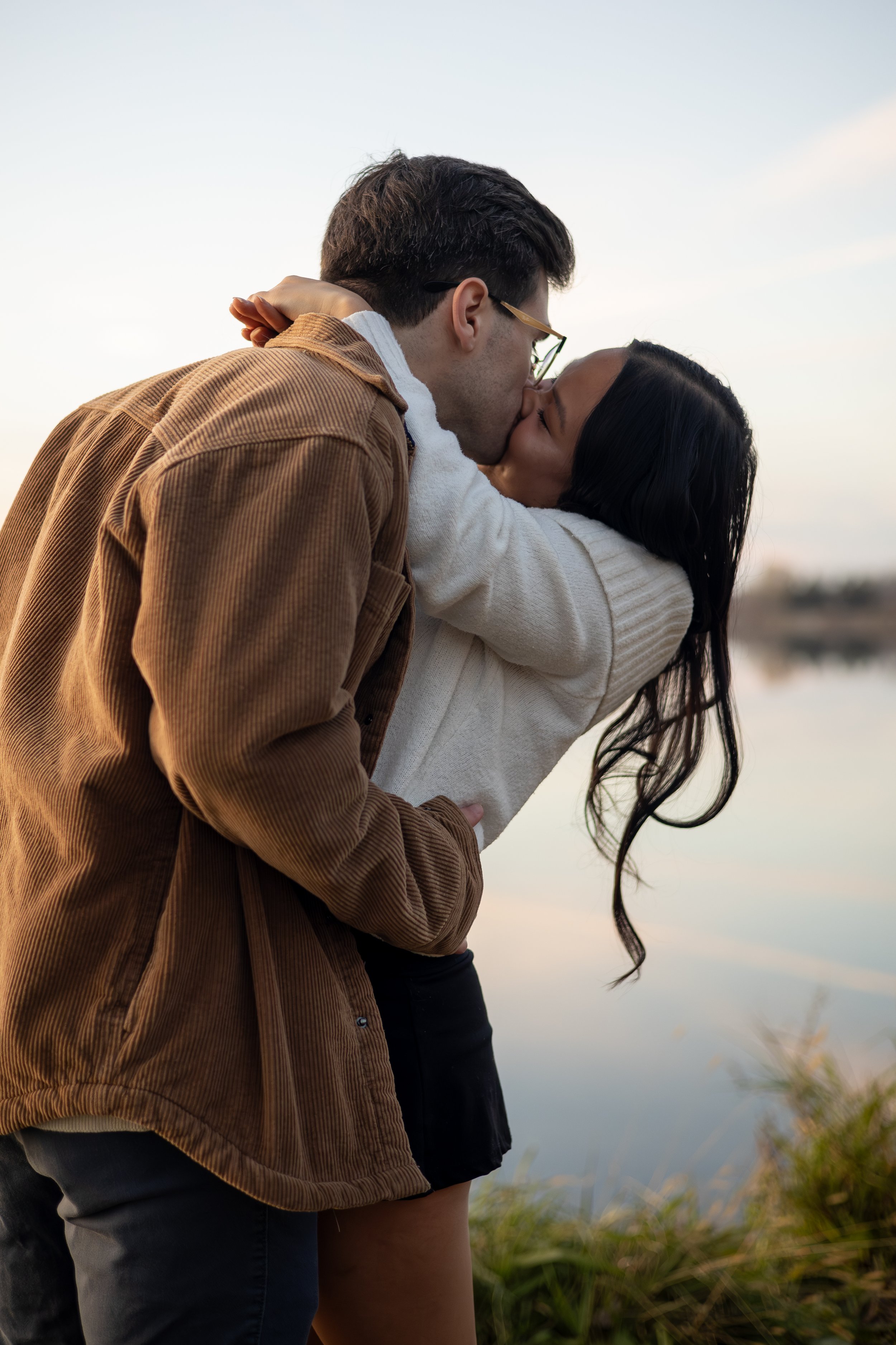 A couple kissing outdoors near a body of water with grass and plants in the foreground, during sunset or dusk.