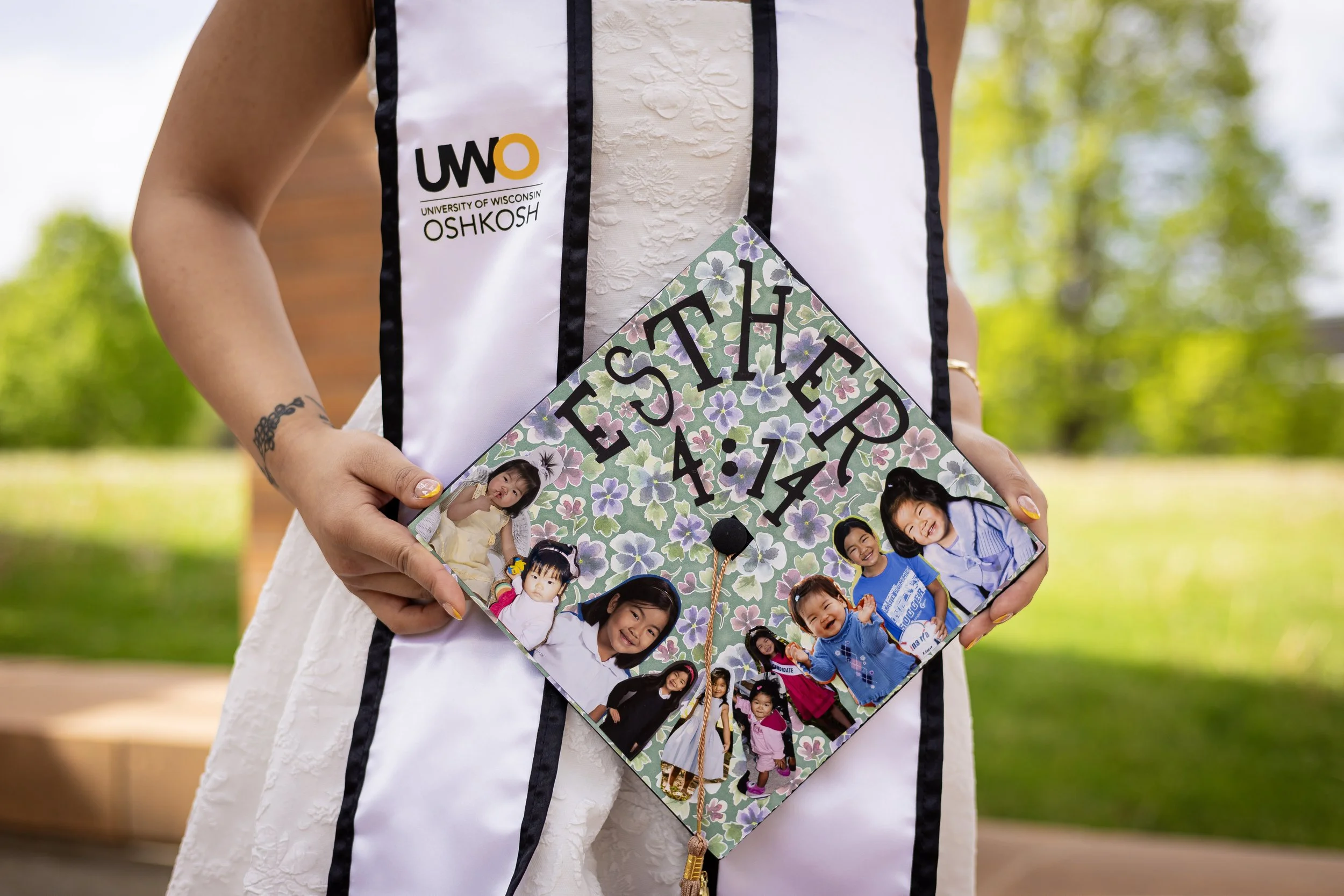 Person holding a graduation cap decorated with photos of children and flowers, wearing a University of Wisconsin Oshkosh sash, outdoors with greenery in the background.