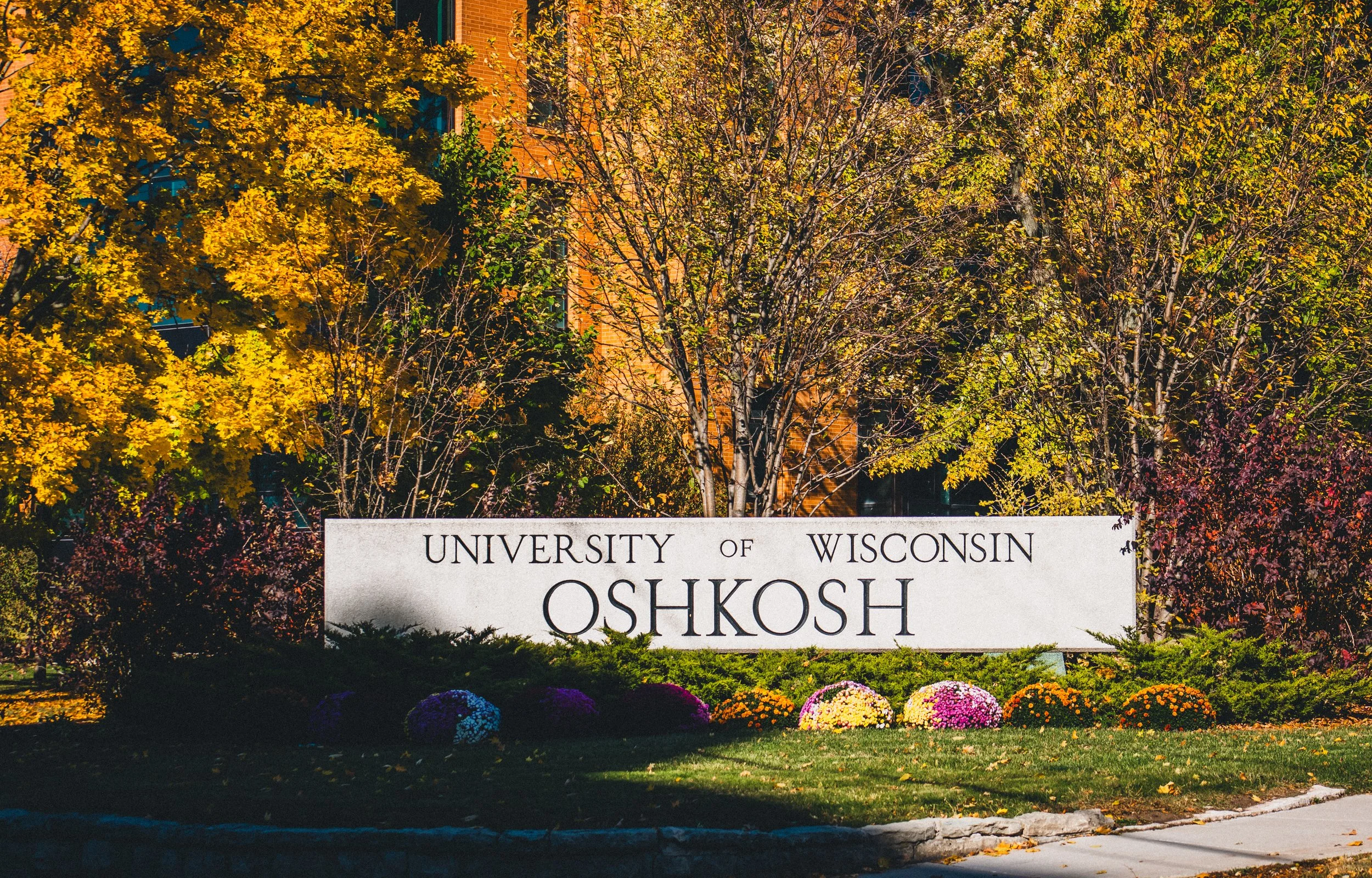 Sign for the University of Wisconsin Oshkosh in front of trees with fall foliage and a building in the background.
