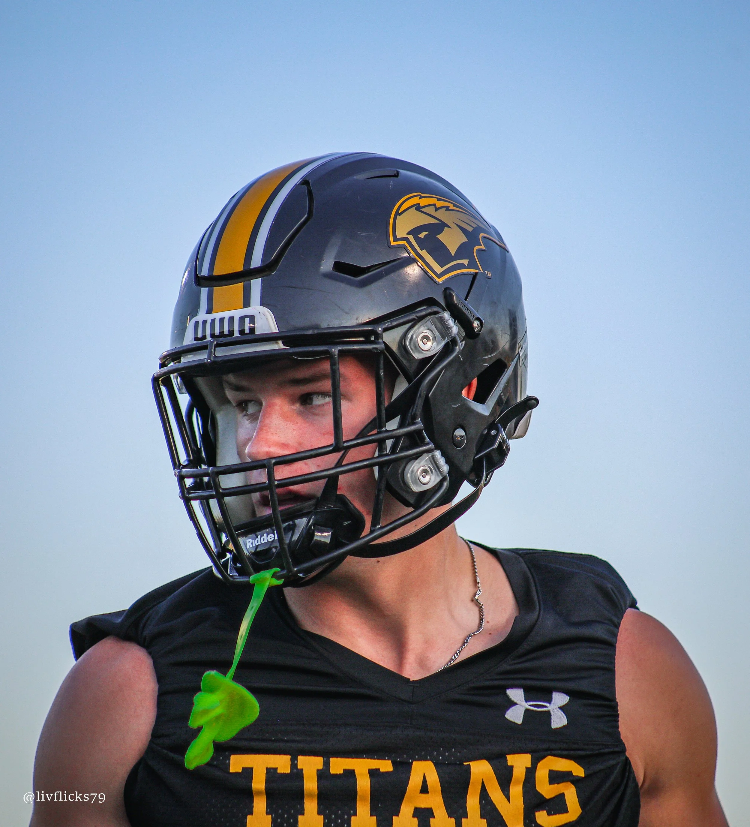 A football player wearing a gray helmet with a Trojan logo, black shirt with orange text, and a chain necklace. The player has a green ribbon on the shoulder, and the background is clear blue sky.