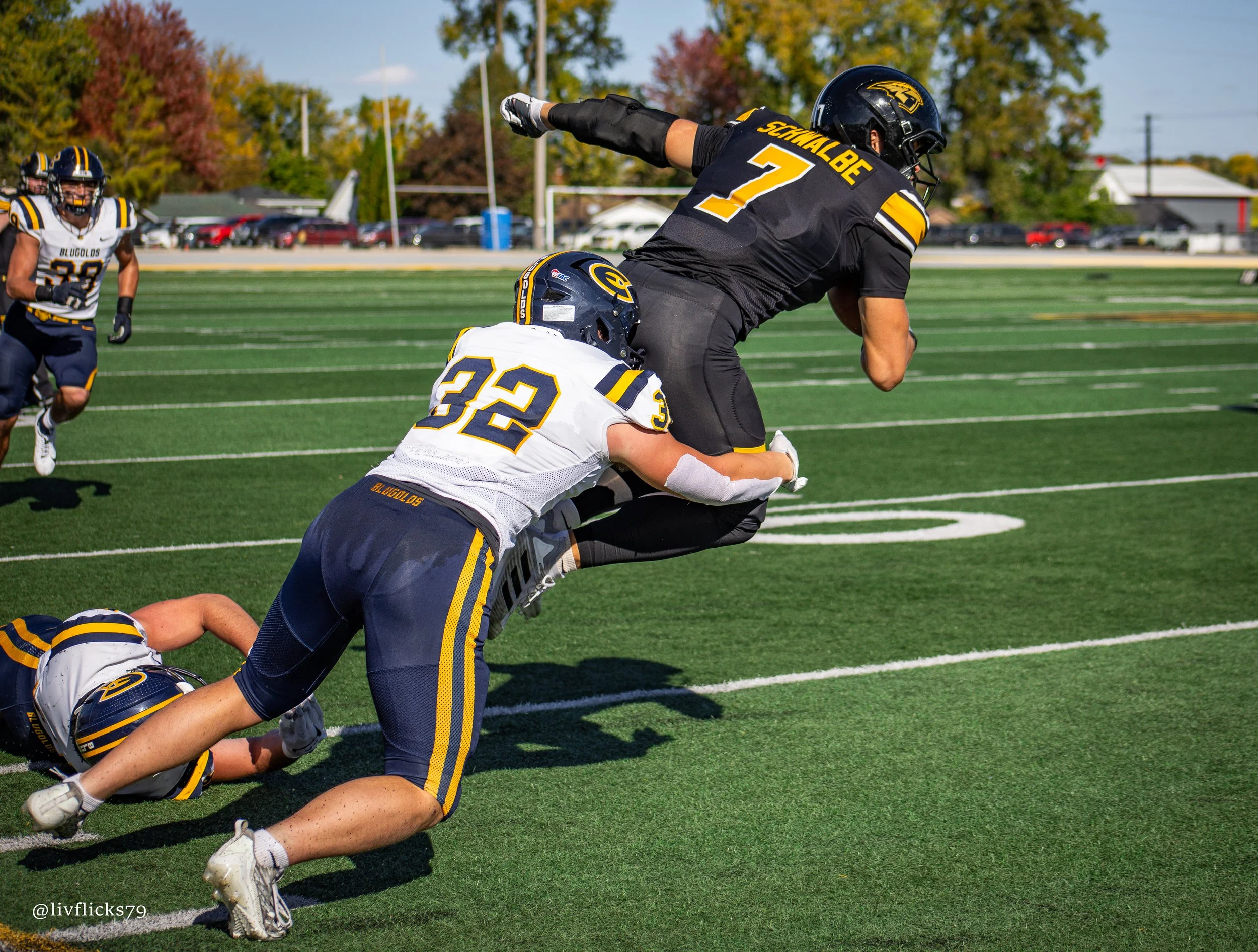 An American football player in black uniform running with the ball, tackled by a player in white uniform on a green football field, with another player in black and two in white chasing in the background.