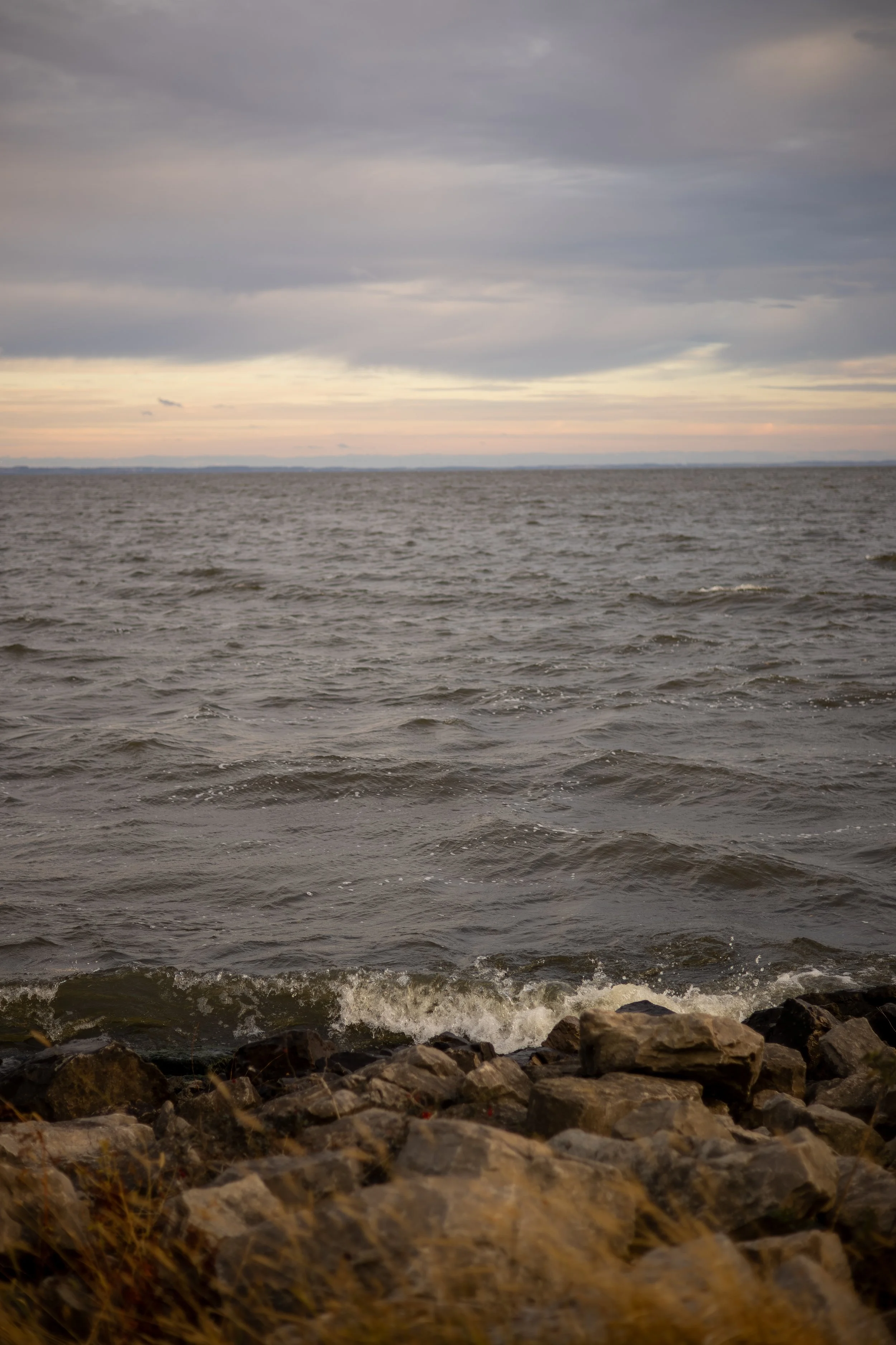 View of a large body of water with small waves, rocky shoreline in the foreground, and a cloudy sky at dusk or dawn in the background.