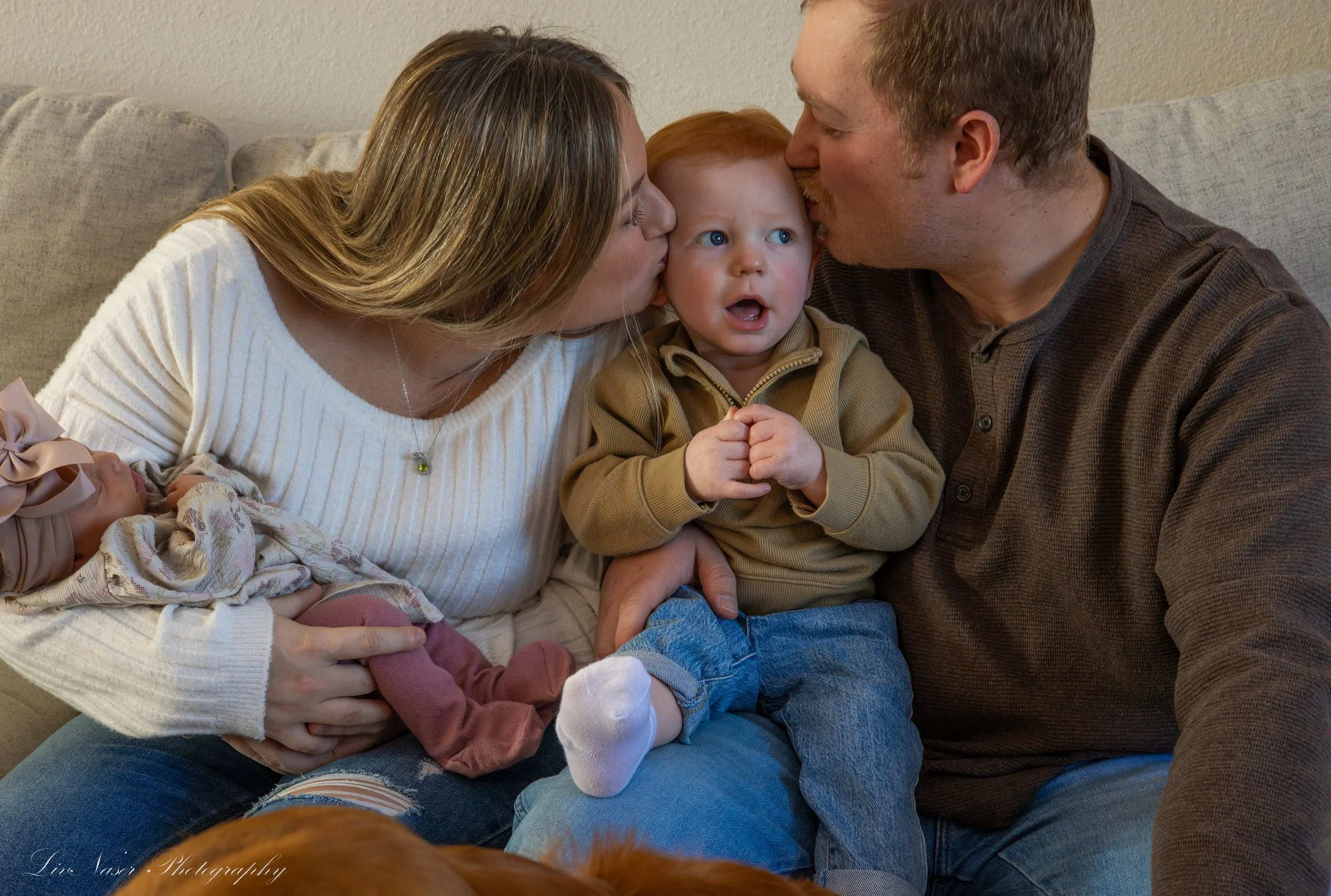 A family of three sitting on a beige couch, sharing kisses with their young child between them. The mother and father are kissing the child's cheeks, and the child has wide eyes and an open mouth, wearing a tan jacket.