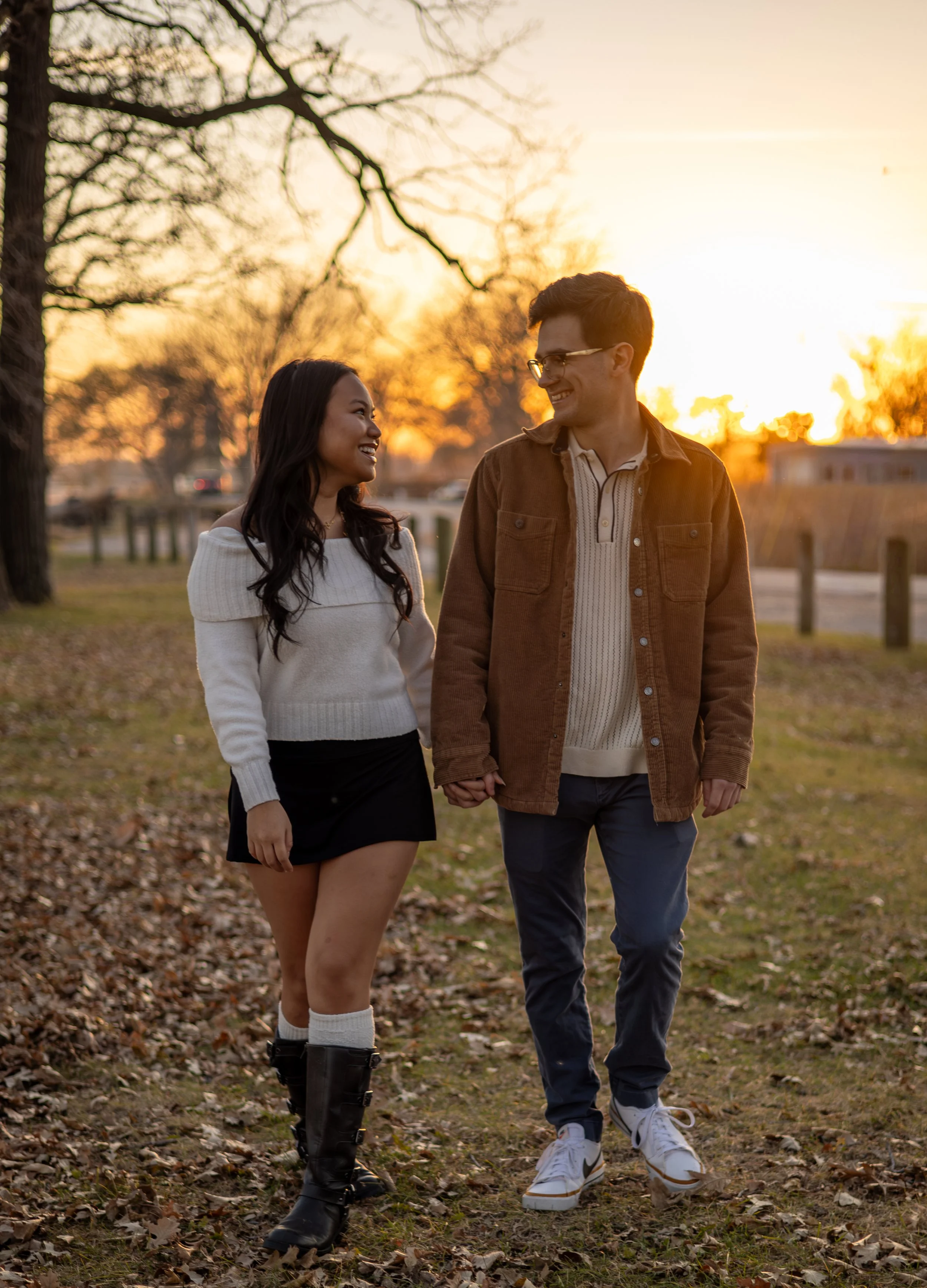 A young couple walking hand-in-hand outdoors during sunset in a park with trees, grass, and fallen leaves, smiling at each other.
