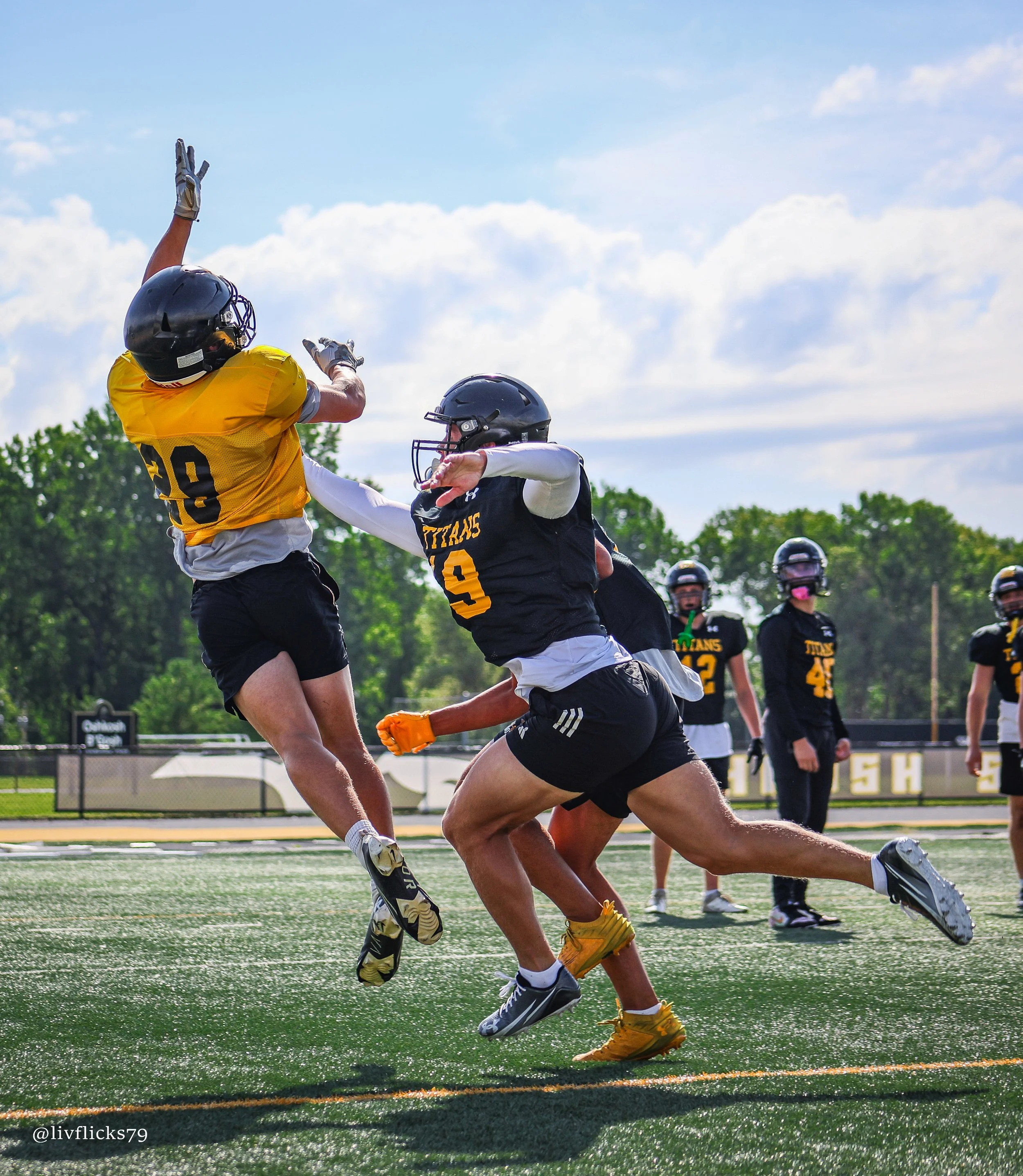 Two football players jump to catch a football during a game or practice on a green field with a fence and trees in the background, under a partly cloudy sky.