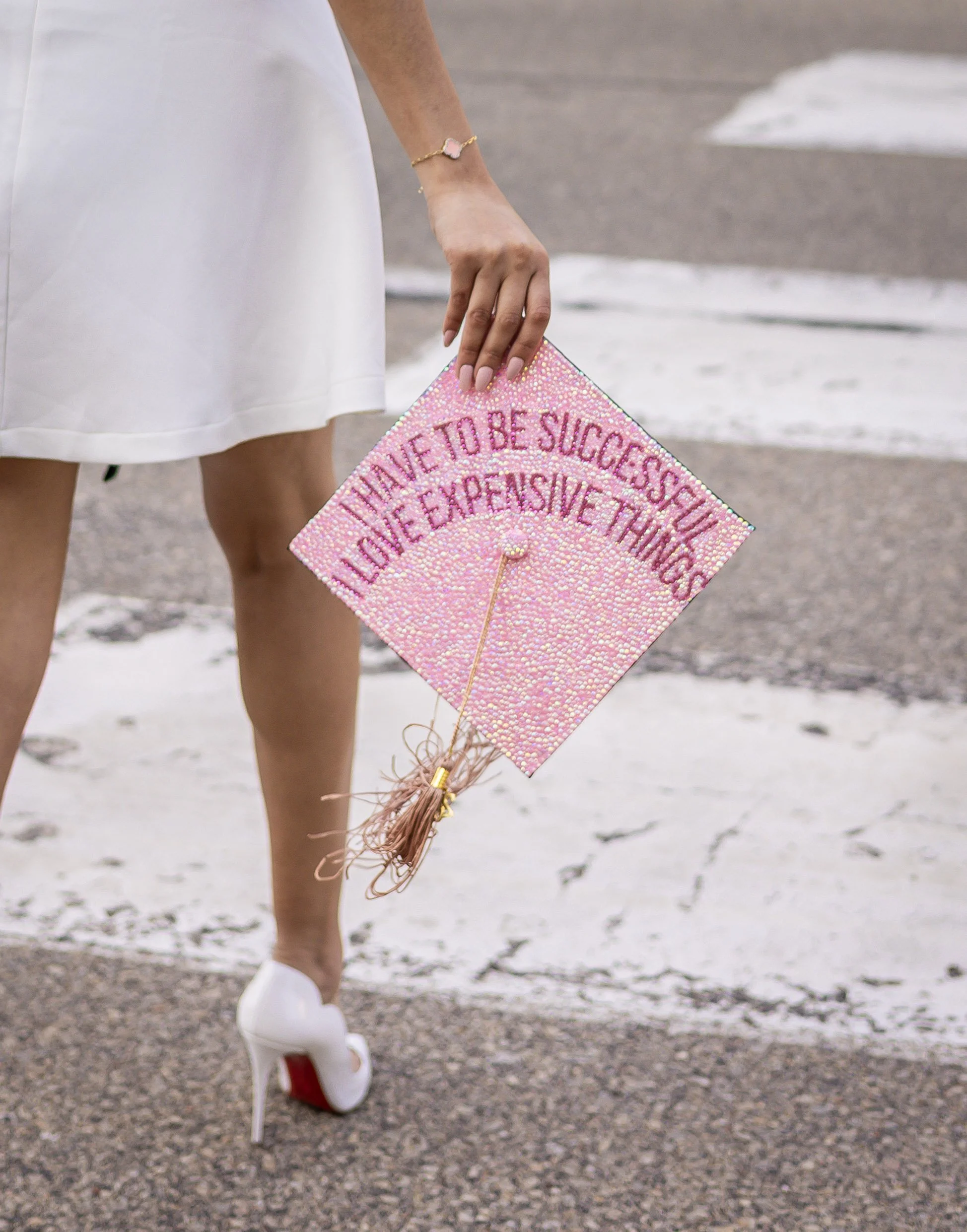 Close-up of a woman in a white dress holding a pink beaded graduation cap that has a message written on it, walking on a street for her graduation photos.