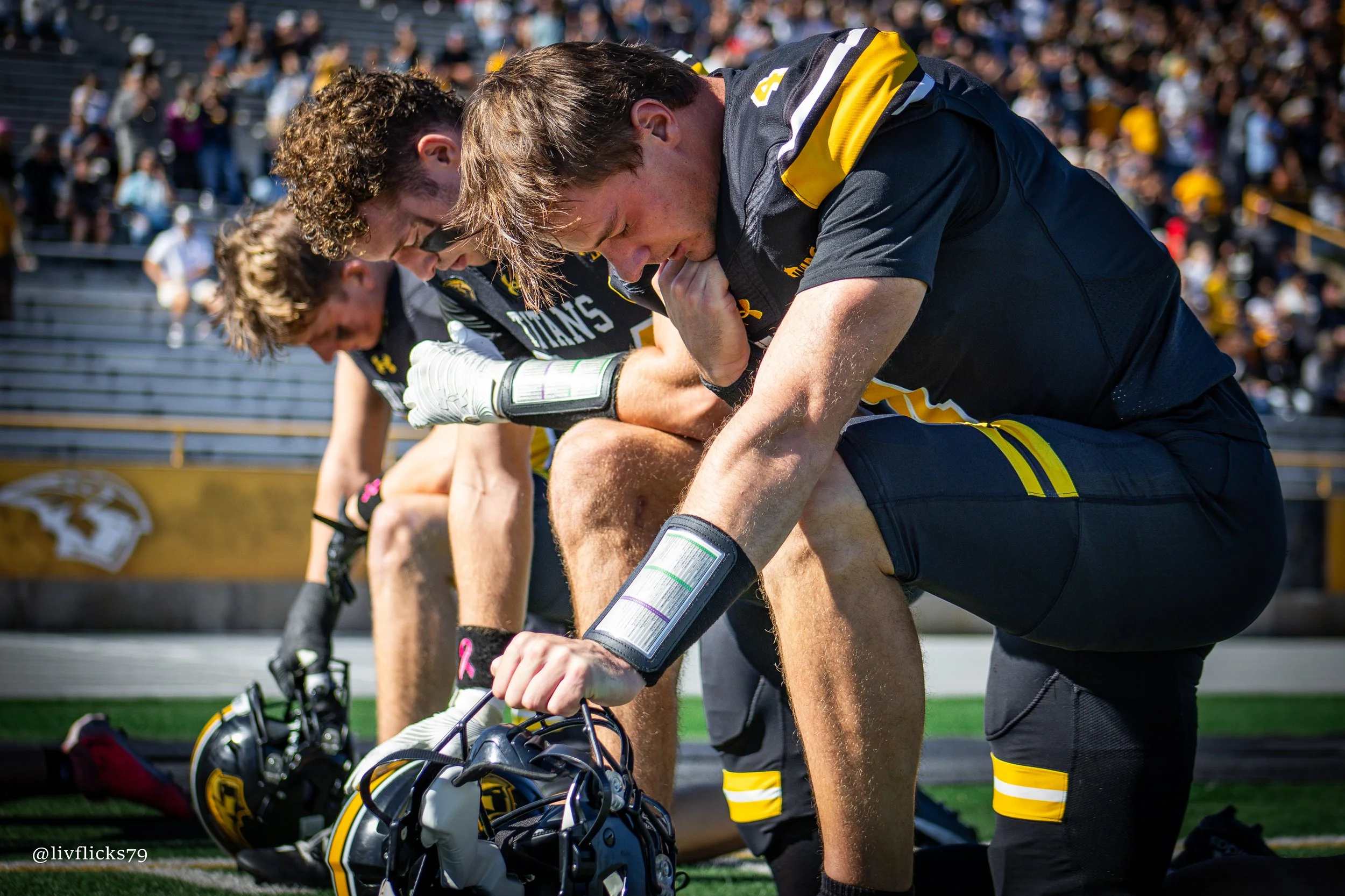 Three football players kneel on the field in prayer with helmets off, hands clasped together, on a sunny day during a game.