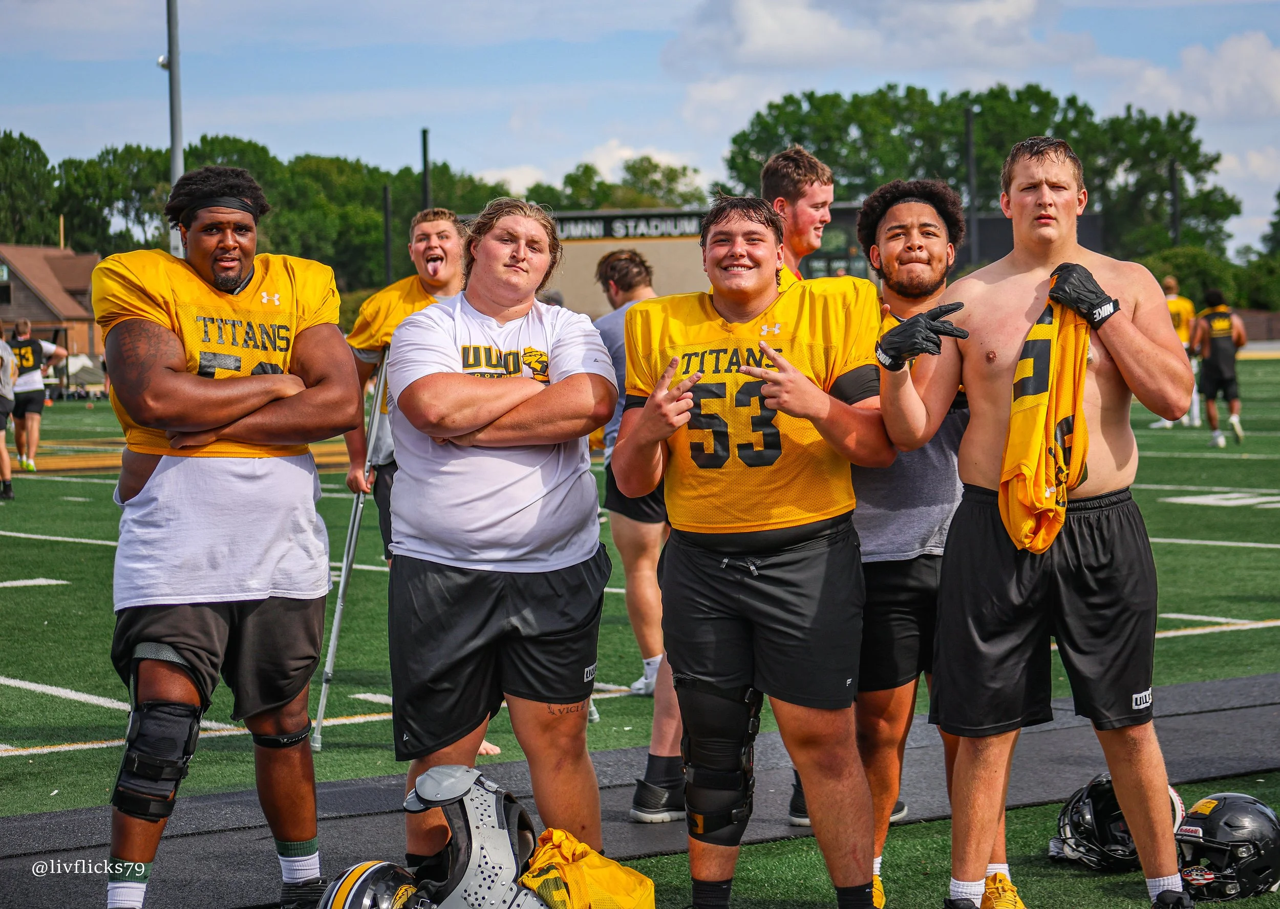 Group of football players on a field, some wearing yellow jerseys, some shirtless, posing and smiling for a photo.