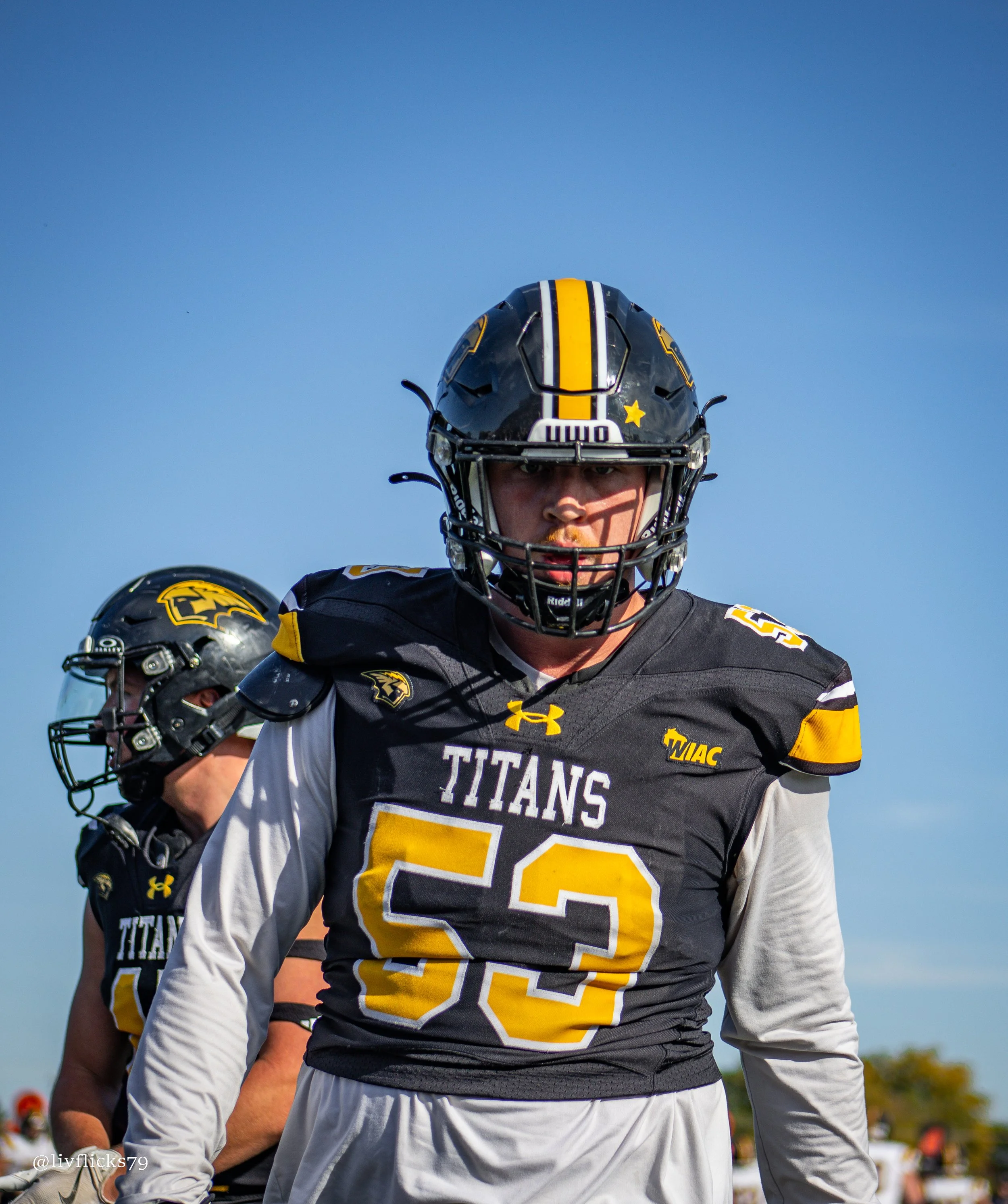 A football player wearing a black and yellow uniform and helmet with the word 'Titan' on the front, standing outdoors under a clear blue sky.