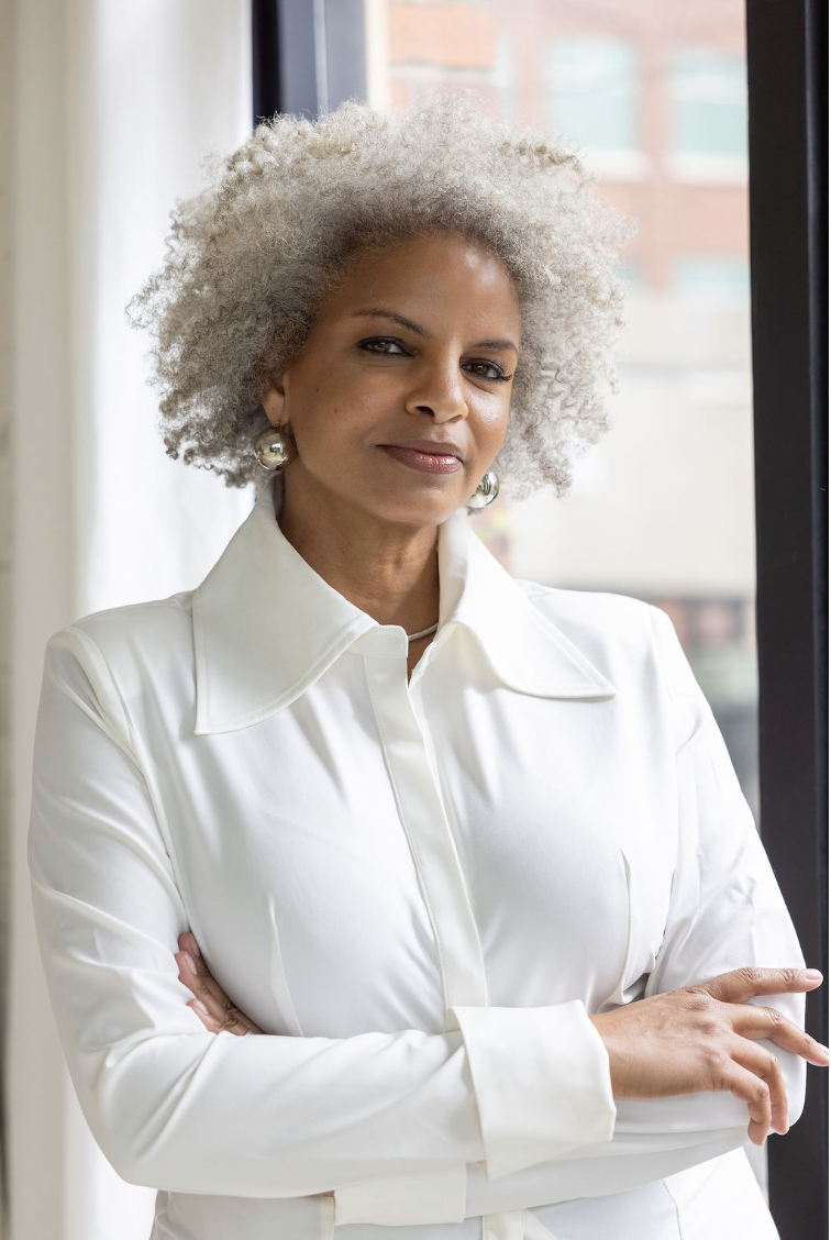 An older woman with curly gray hair wearing a white blouse and silver earrings standing with arms crossed near a window.