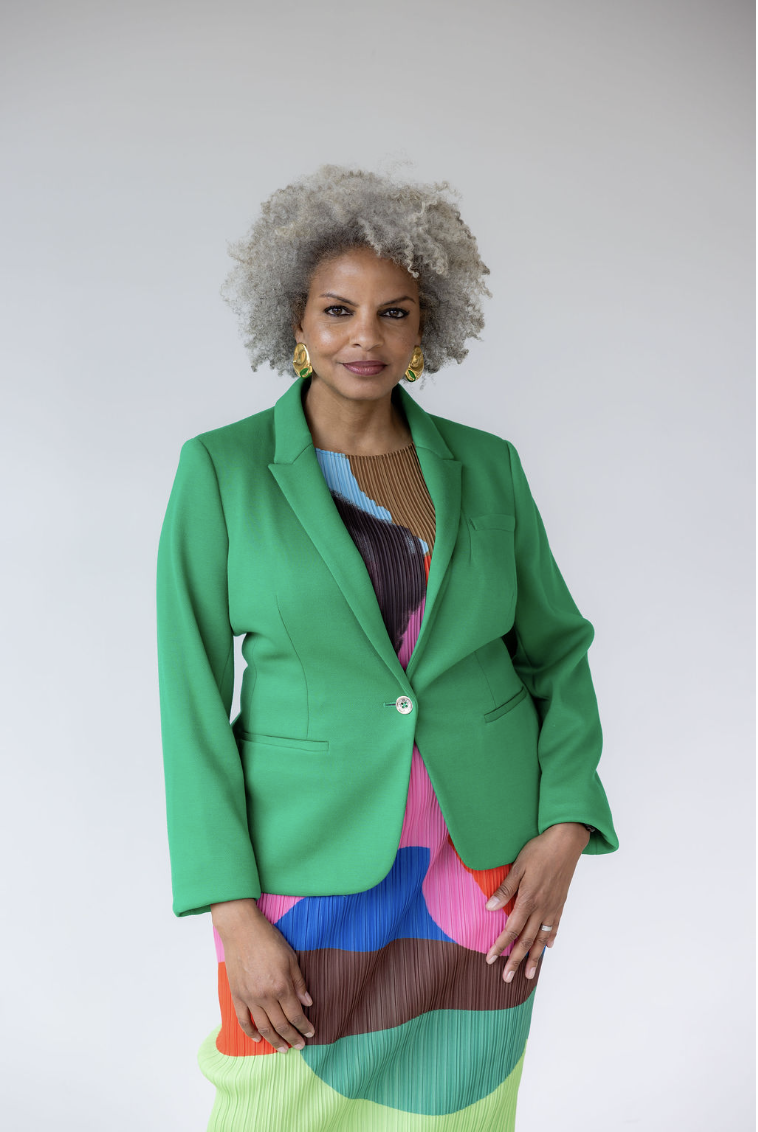 A woman with curly gray hair wearing a bright green blazer over a colorful patterned dress, standing against a plain white background.