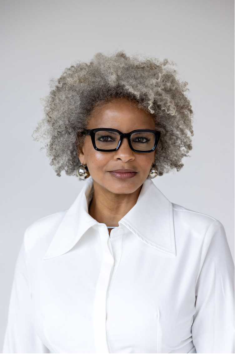 A stylish middle-aged woman with curly gray hair wearing black glasses, silver earrings, and a white collared shirt against a plain background.