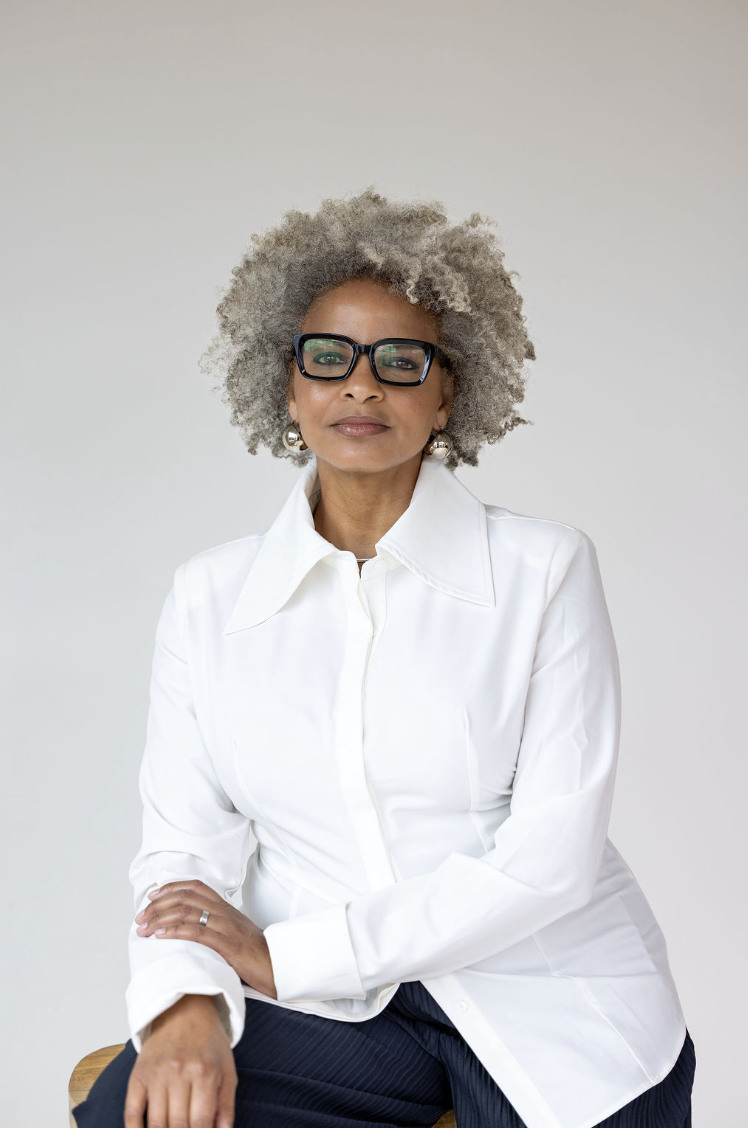 A woman with gray curly hair wearing glasses, a white button-up shirt, and dark pants, sitting on a wooden stool against a plain light background.