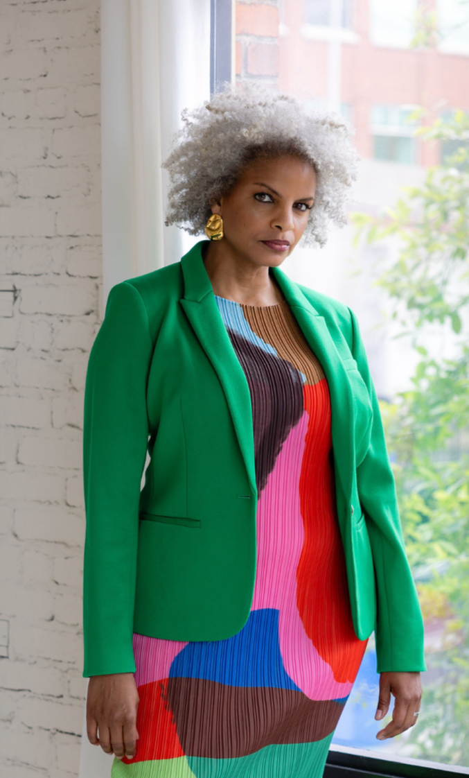 A confident woman with curly gray hair wearing a bright green blazer and a colorful, abstract patterned dress, standing near a window with a brick wall and greenery outside.